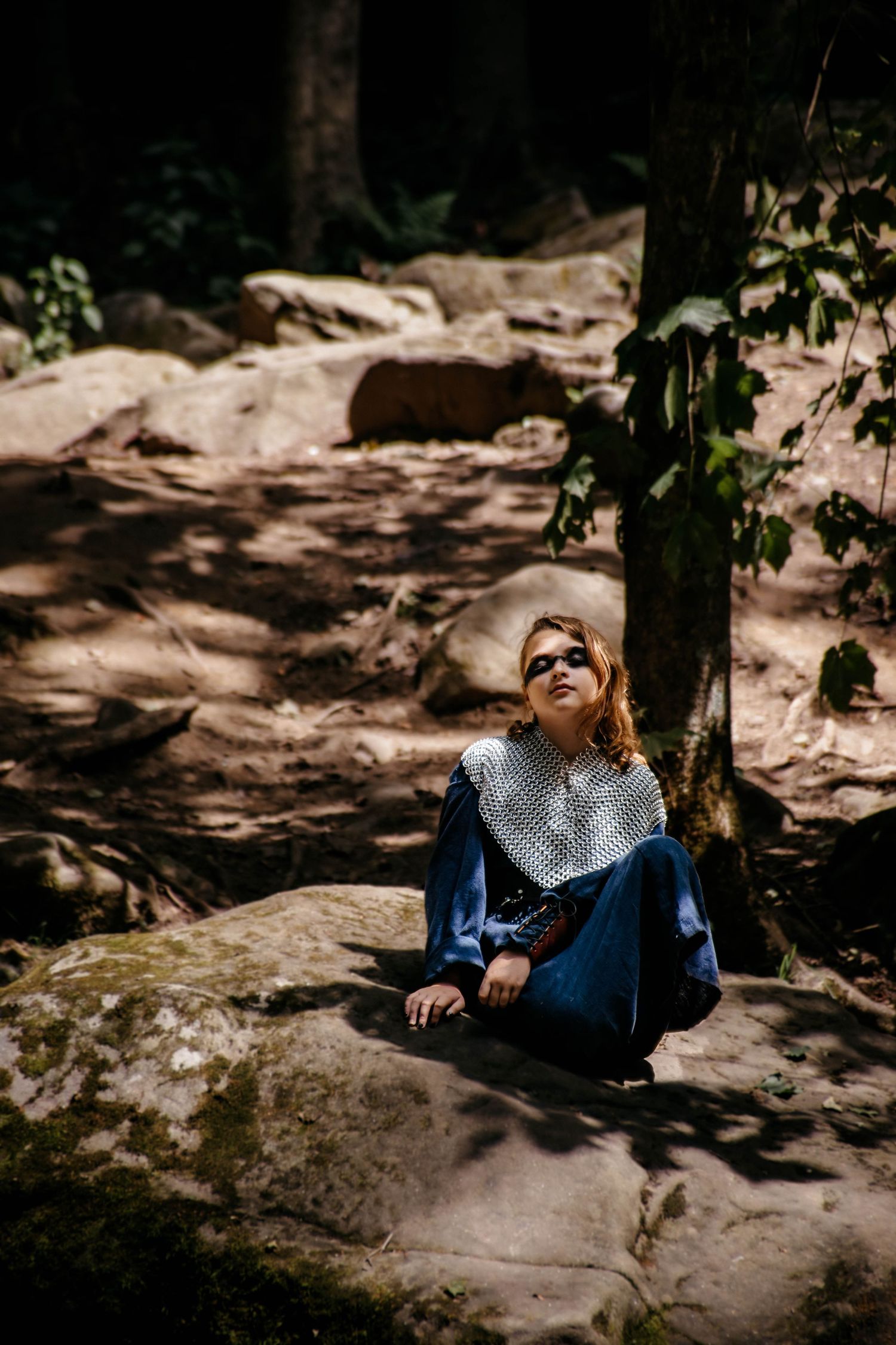 A person in a polka dot top and jeans reclines on a large rock in dappled sunlight beneath trees.