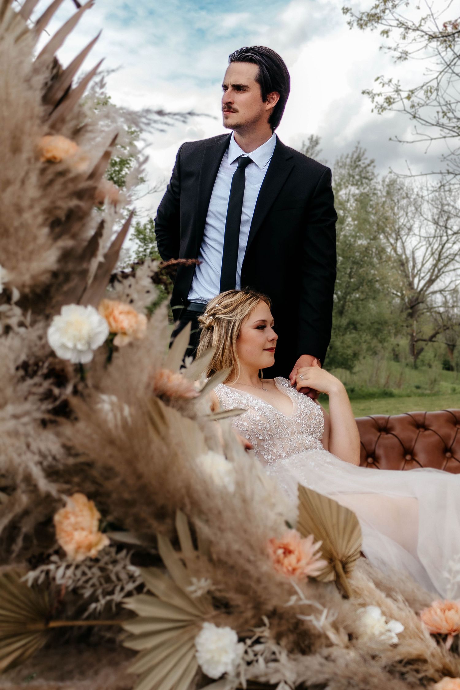 A wedding couple poses near dried pampas grass and floral arrangements on a vintage brown leather couch outdoors.