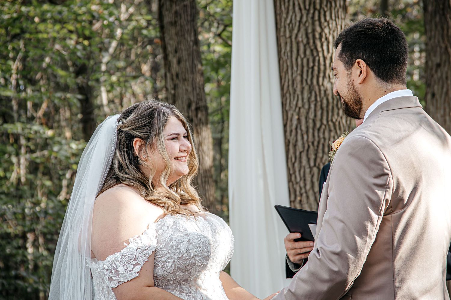 A couple shares a joyful moment together during an outdoor wedding ceremony beneath tall trees and white drapes.