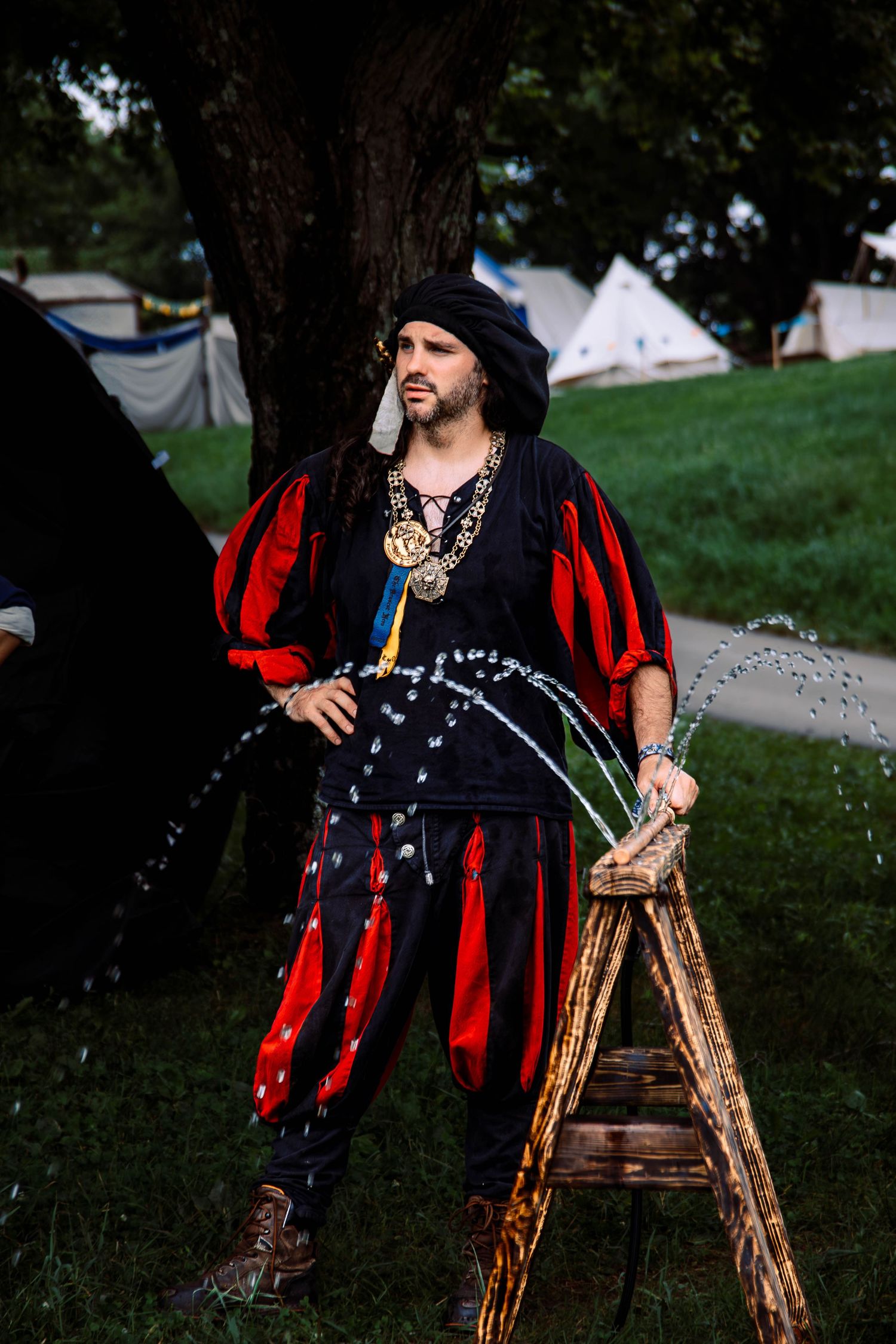 A performer in medieval Renaissance faire costume with red and black striped clothing poses next to a wooden ladder outdoors.