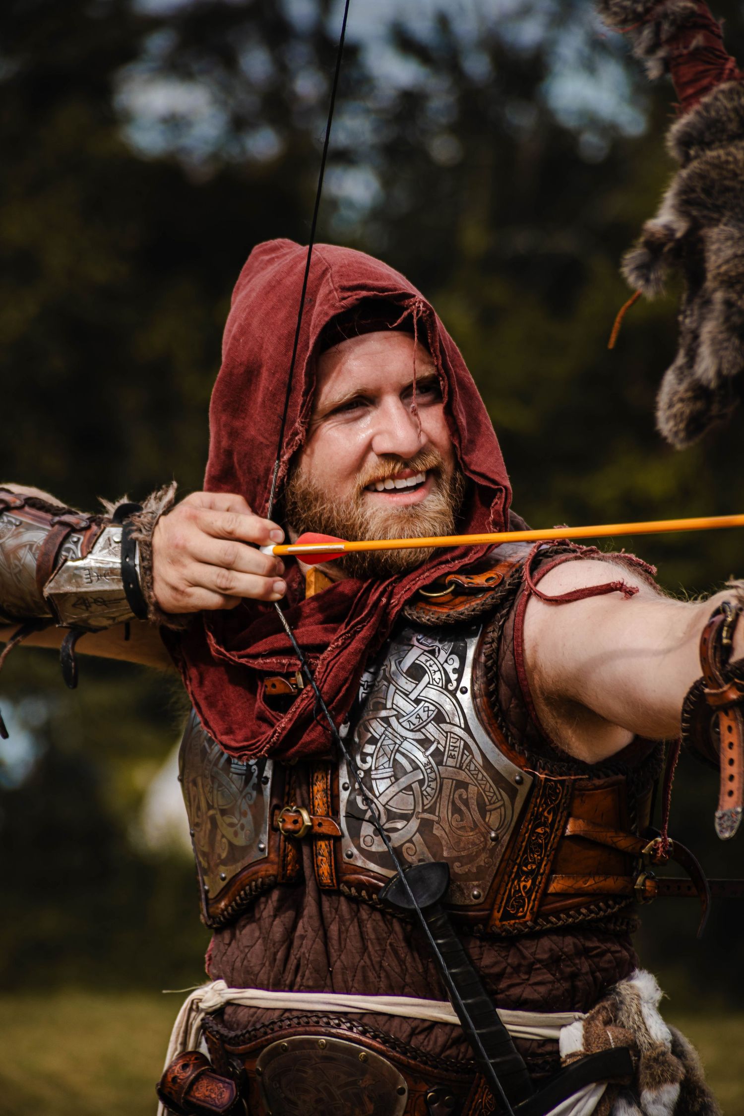 Medieval archer in leather armor and red hood draws a wooden bow while practicing archery in an outdoor setting.