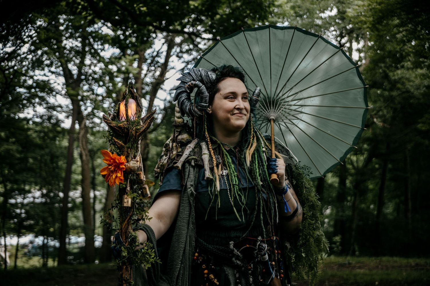 Woman in fantasy forest costume with parasol, staff, and floral accents poses outdoors among trees.