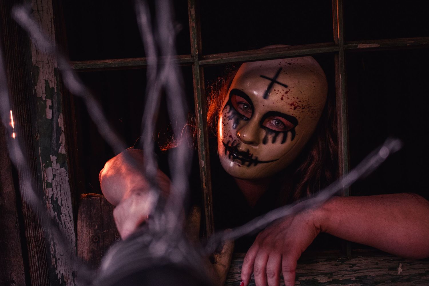 Creepy Halloween costume with painted doll face makeup, cross on forehead, peering through dark wooden window with cobwebs.