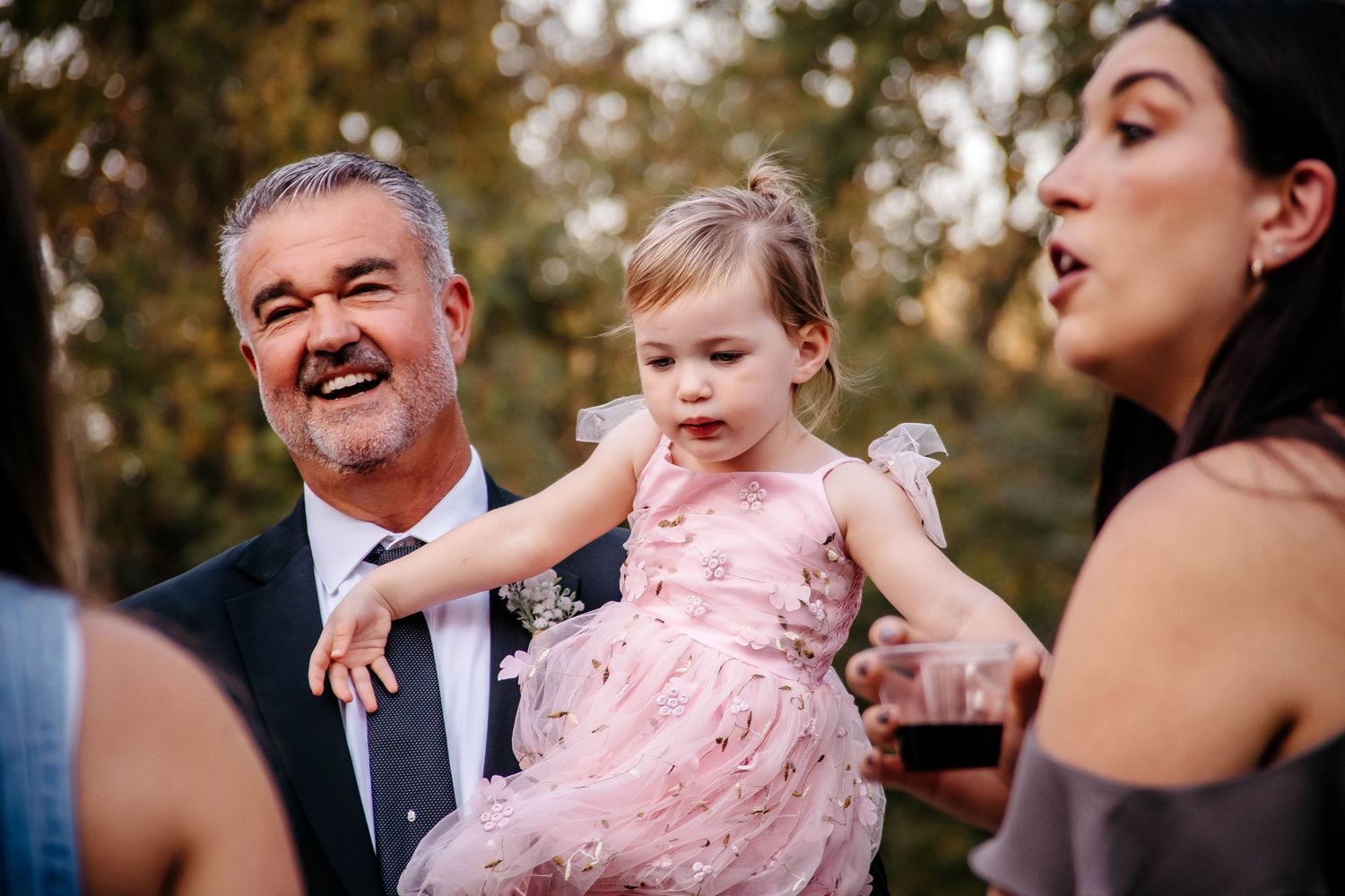 Man in suit holding toddler girl in pink dress at outdoor wedding celebration with guests nearby.