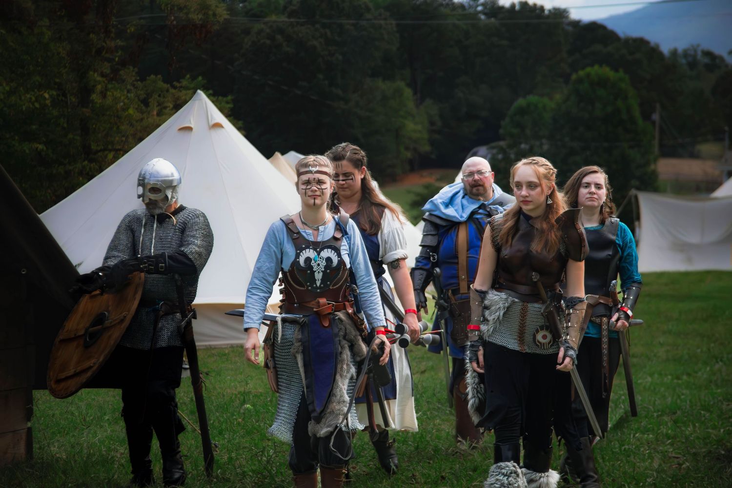 A group of medieval reenactors in period costumes walking across a field with white tents in the background.