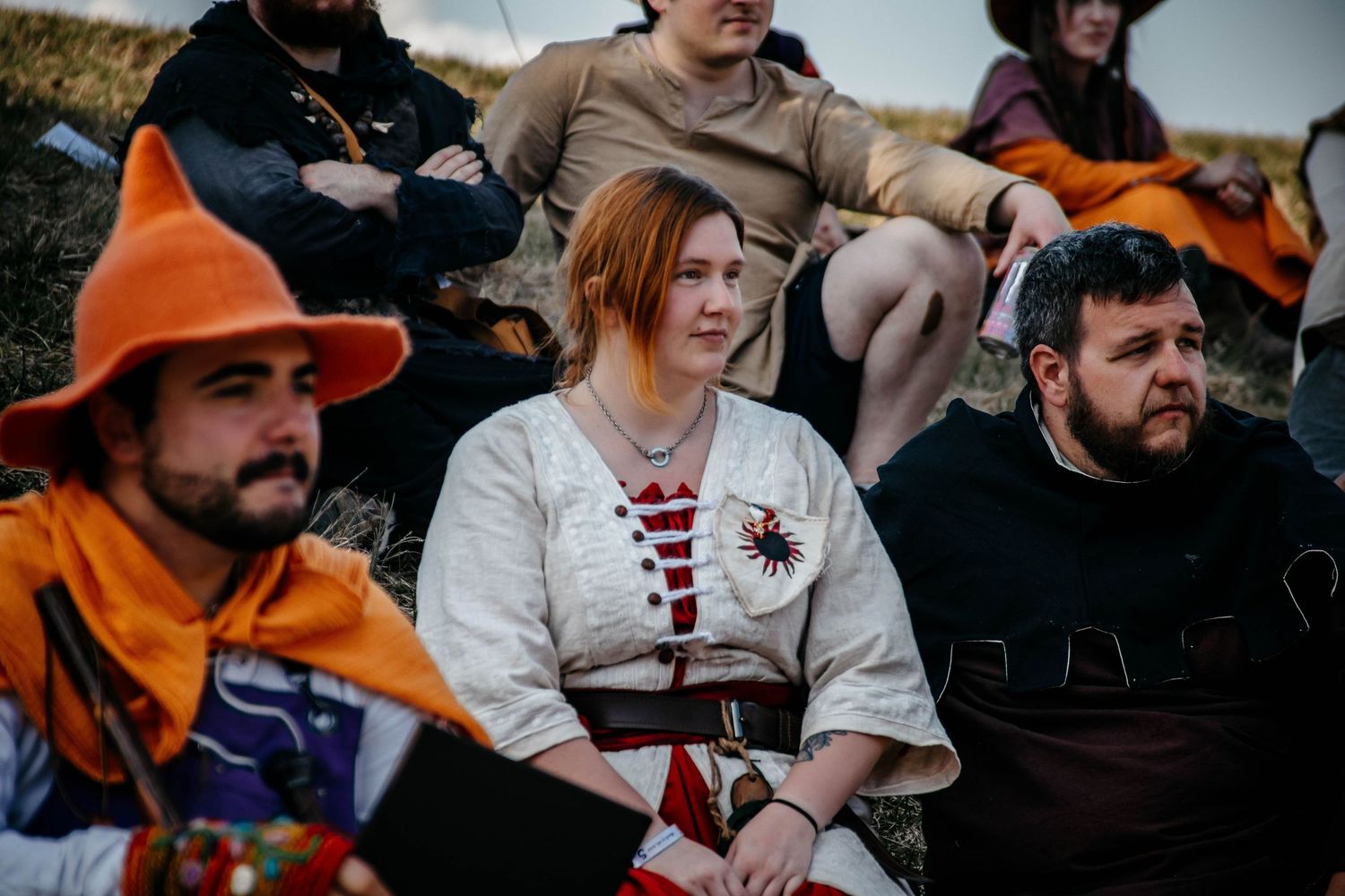 Medieval reenactors in period costumes sit together during an outdoor gathering with orange and brown attire.