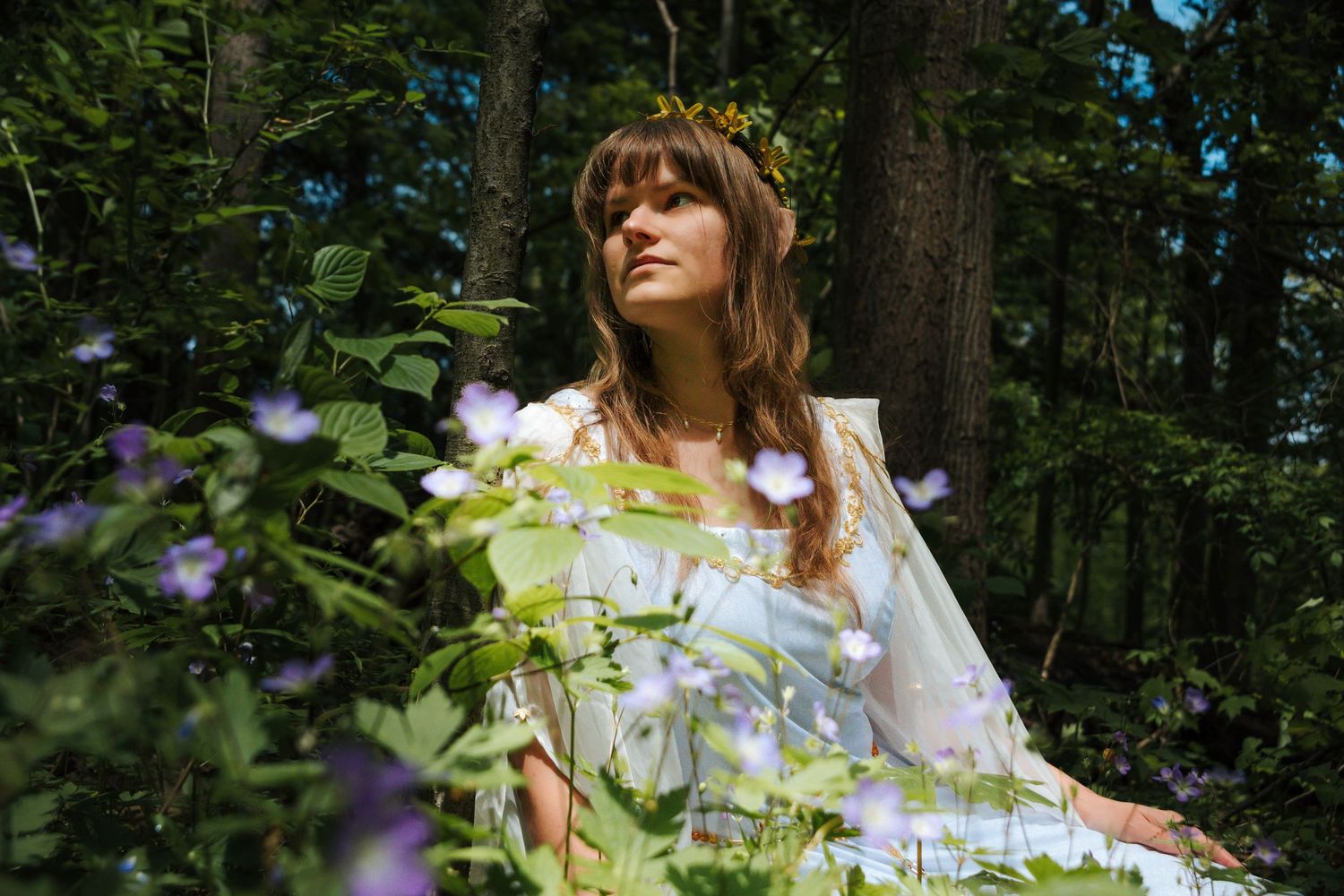 A person in a white flowy dress poses gracefully among wildflowers and trees in a sunlit forest setting.