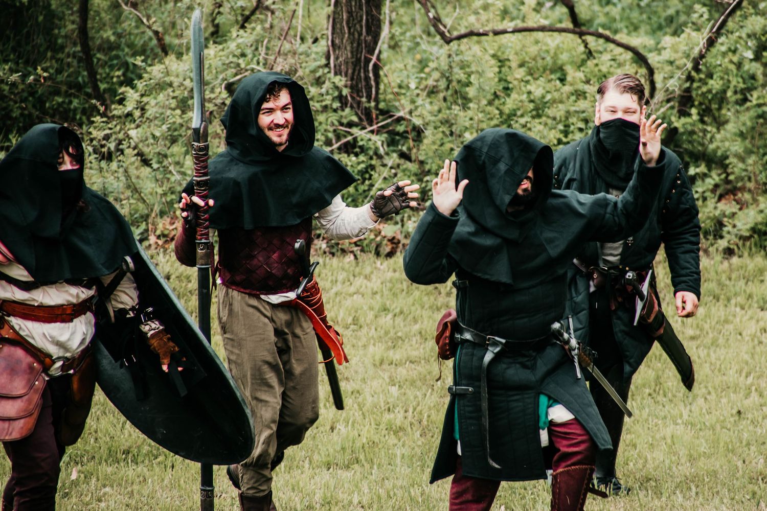 Medieval cosplay enthusiasts in period garments engage in mock combat with swords and shields in a grassy woodland area.