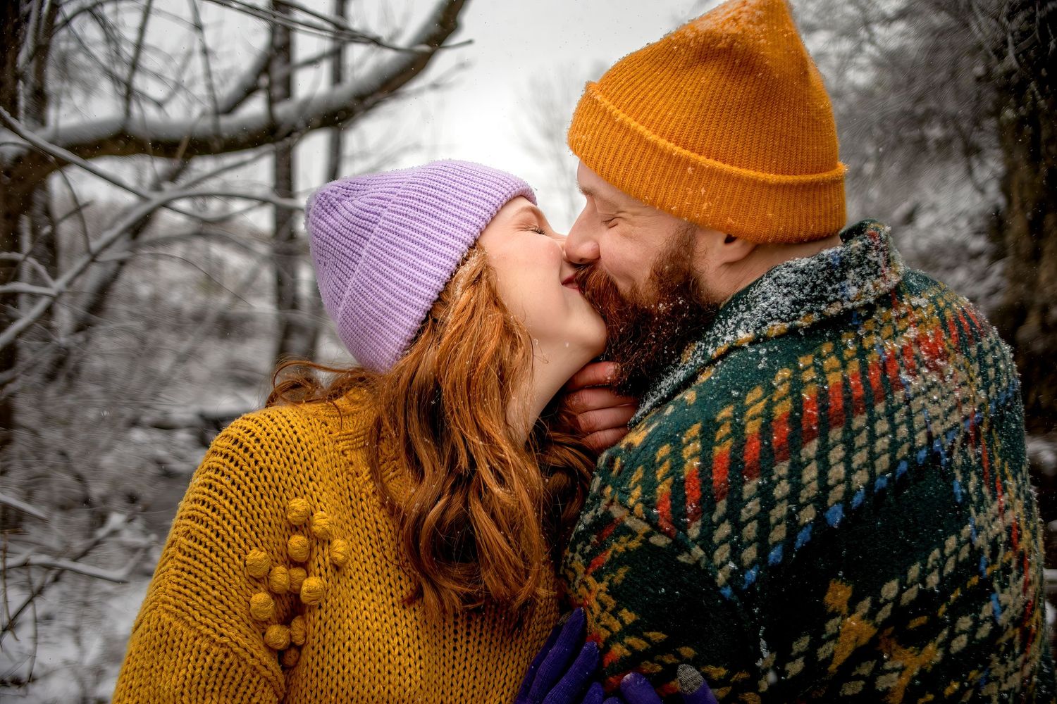 A romantic winter scene shows a couple in knit hats and sweaters embracing in a snowy forest setting.