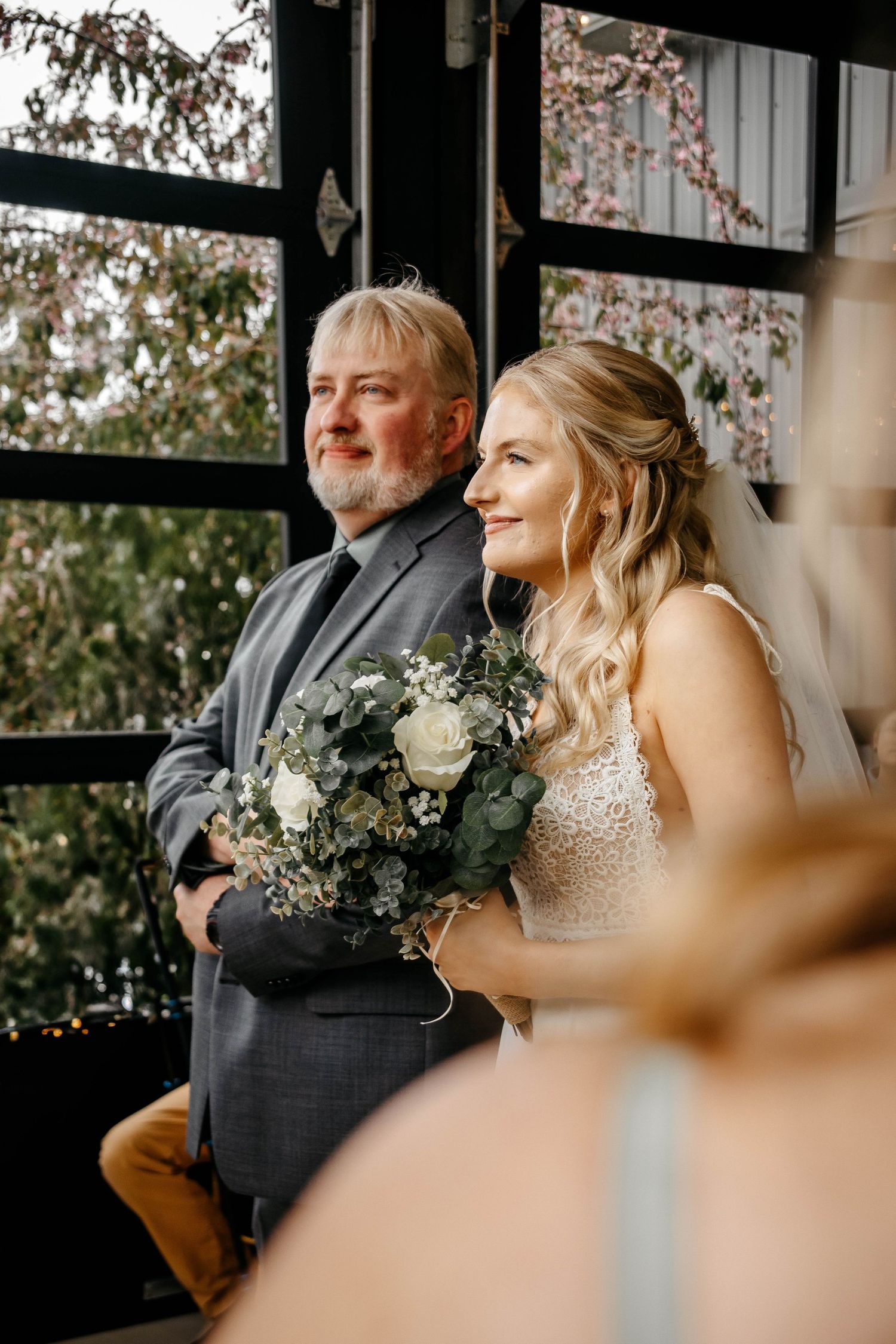 Bride and father share tender moment before wedding ceremony while sitting by large windows with natural light.