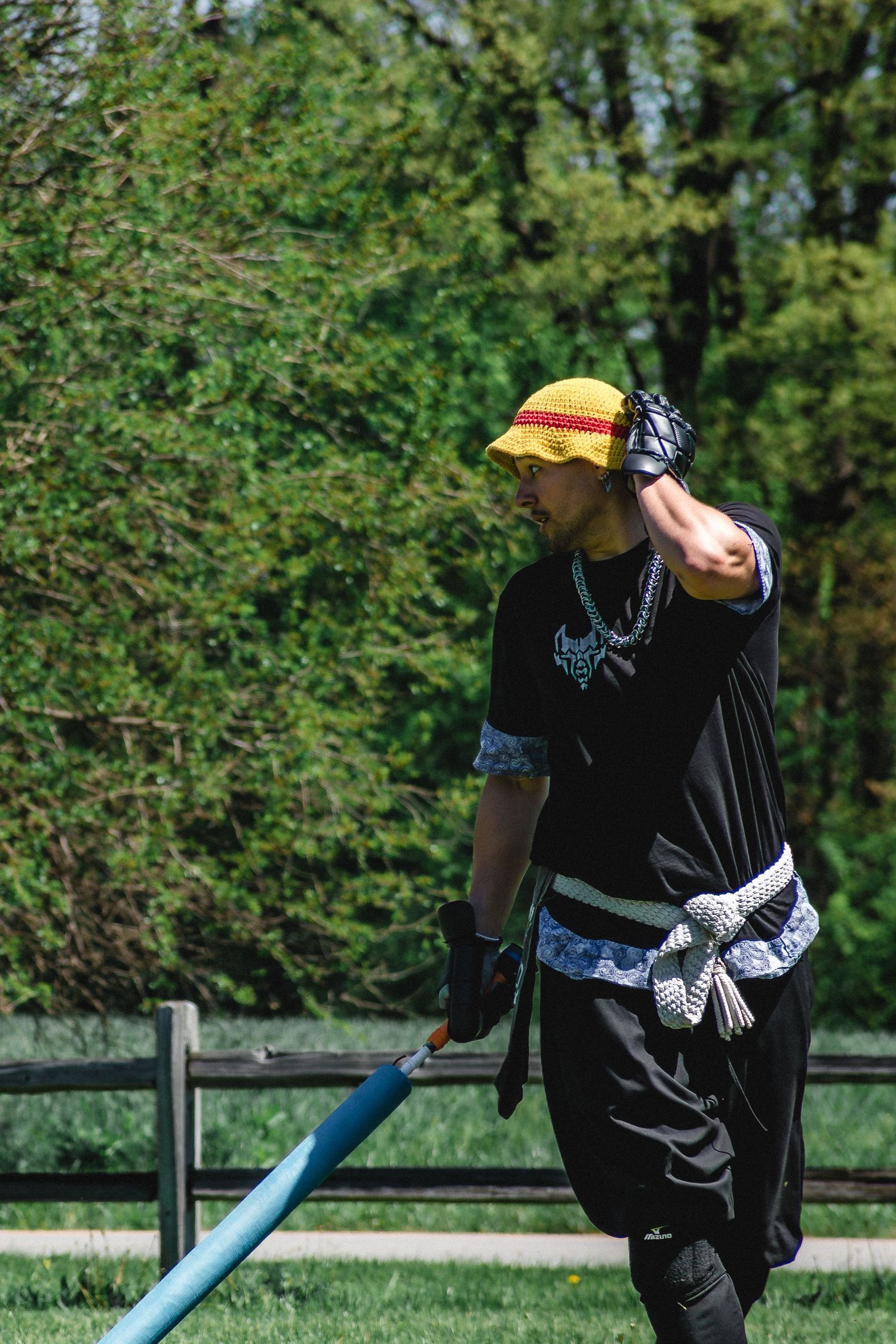 Man in black outfit and yellow hat holding a blue pole outdoors in a park setting.