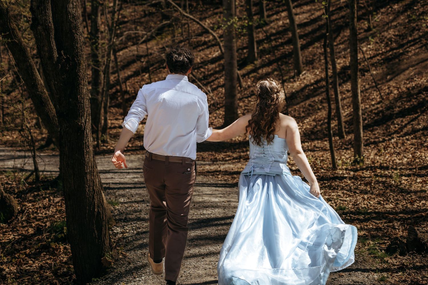 Couple in wedding attire walking hand-in-hand along a forest path, bride in light blue gown.