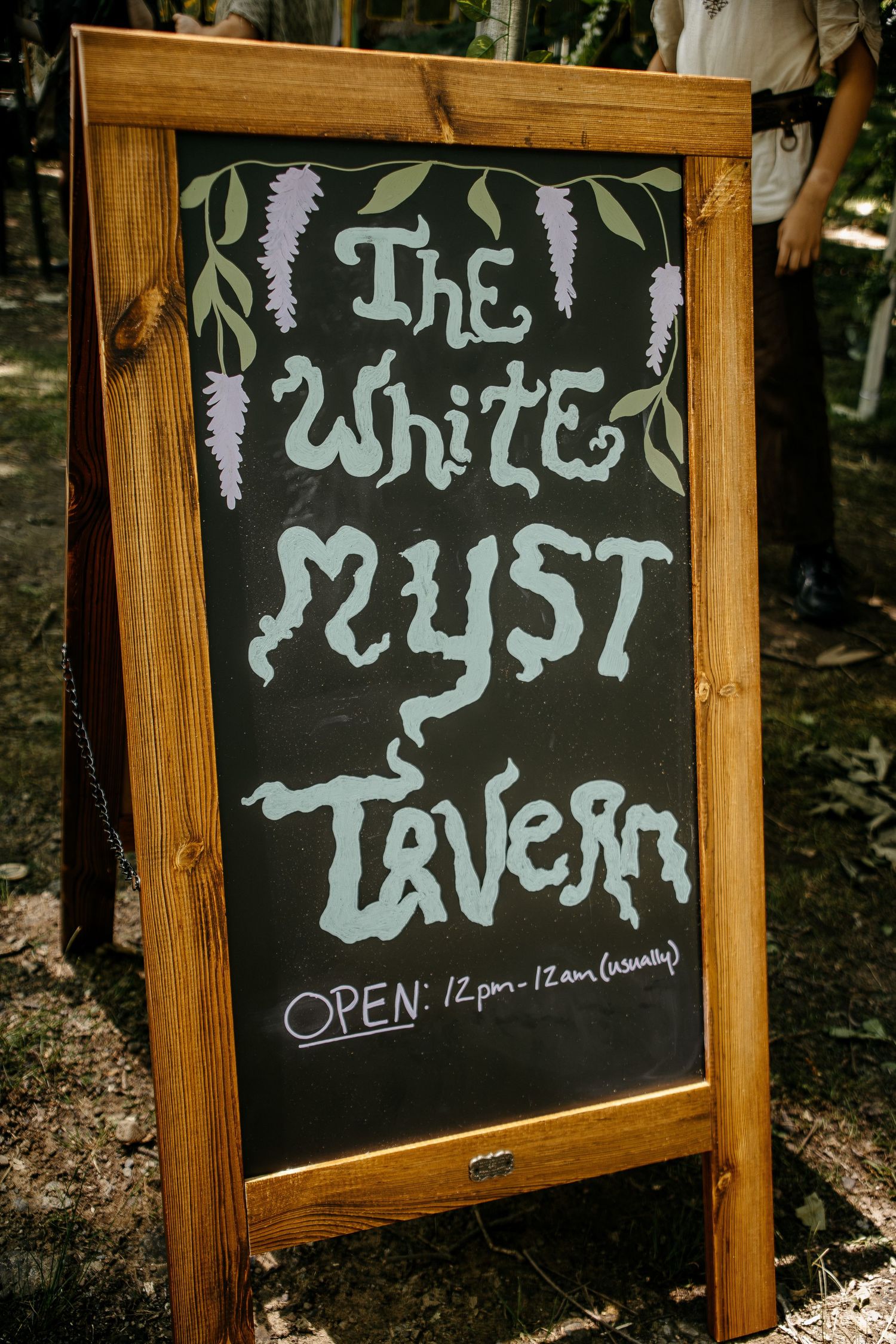 Wooden chalkboard sign for The White Myst Tavern, open 12pm-12am, decorated with purple wisteria flowers.