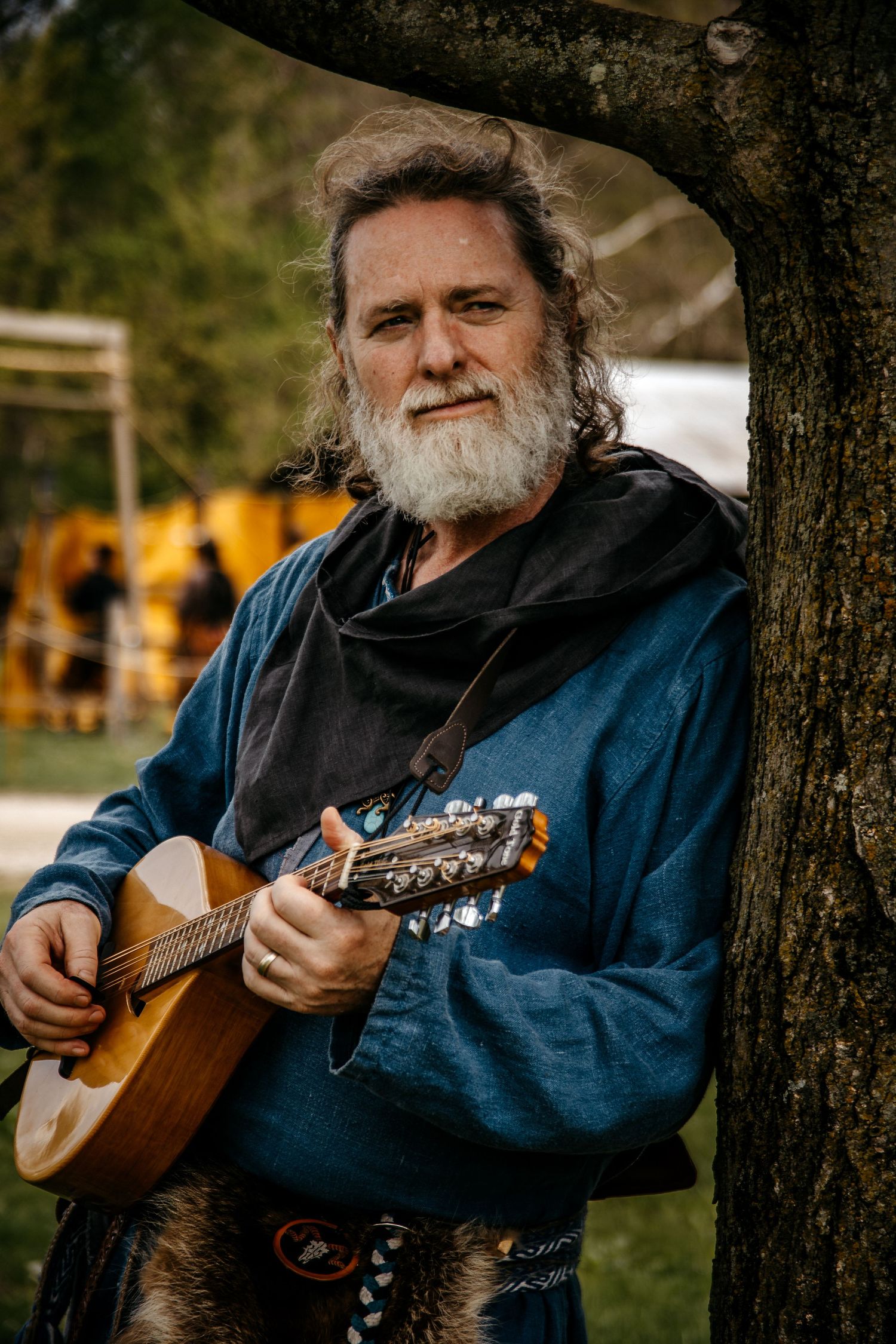 Bearded man with long hair playing a mandolin outdoors, wearing medieval-style blue tunic leaning against a tree.