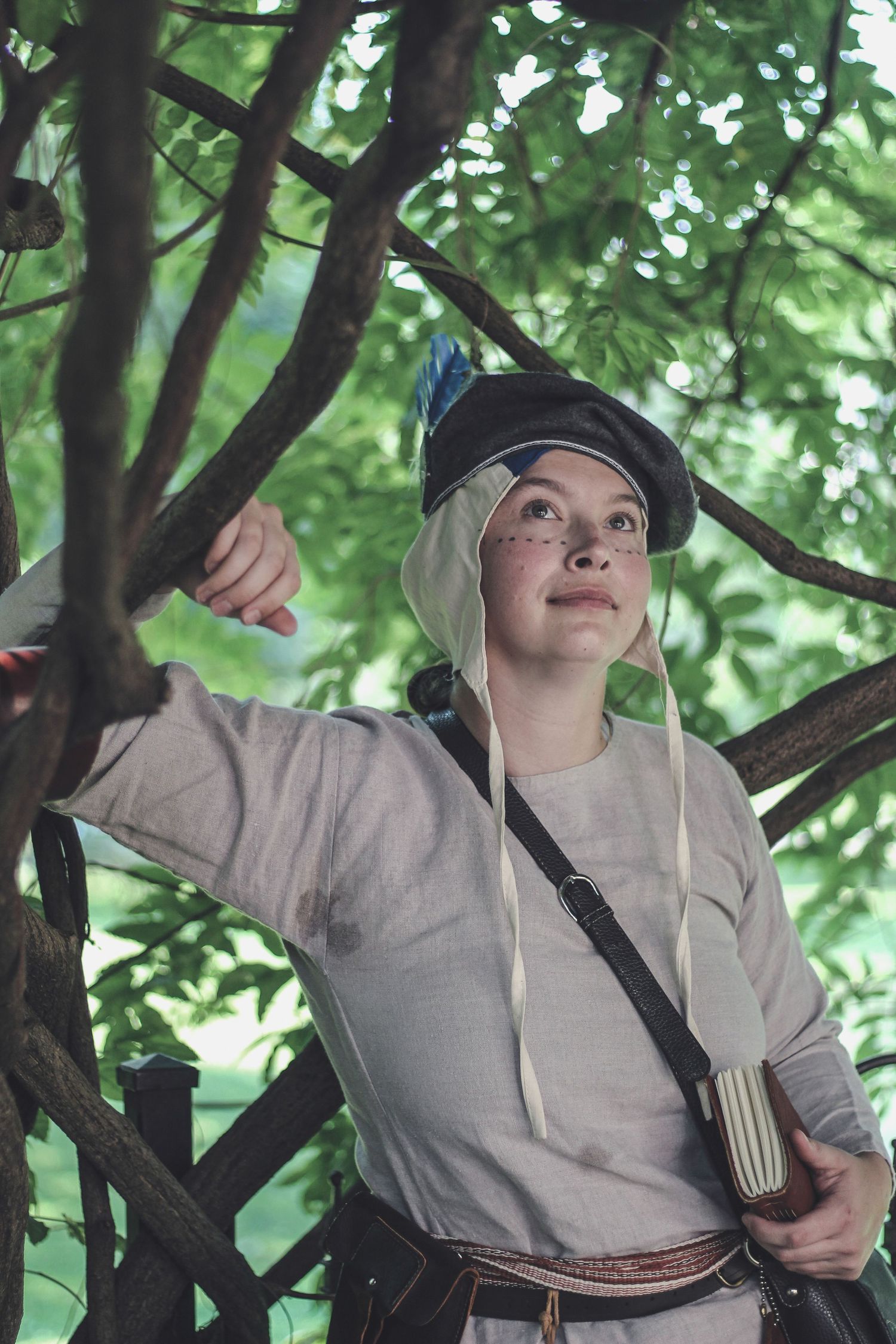 A person in casual clothing and a cap stands among tree branches while holding books and wearing earbuds.