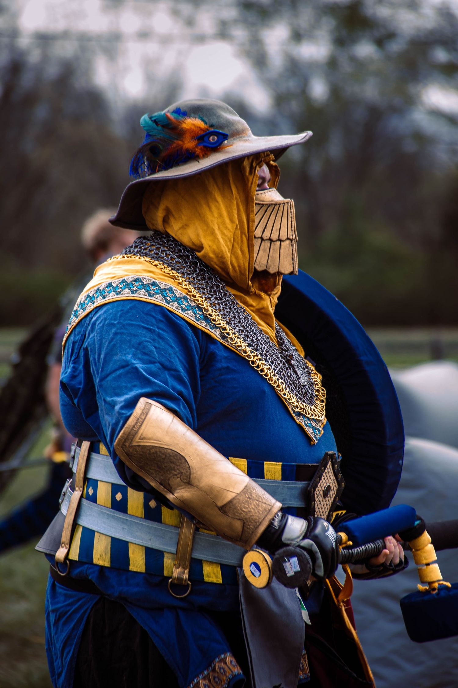 Medieval cosplayer in ornate blue and gold costume with hat and armor at renaissance faire event.