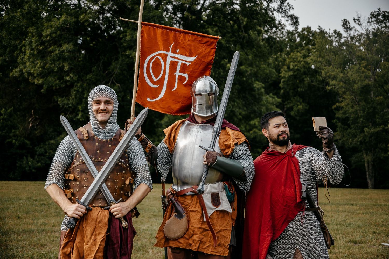 Three men in medieval knight costumes with swords, armor, and a banner pose for a selfie outdoors.