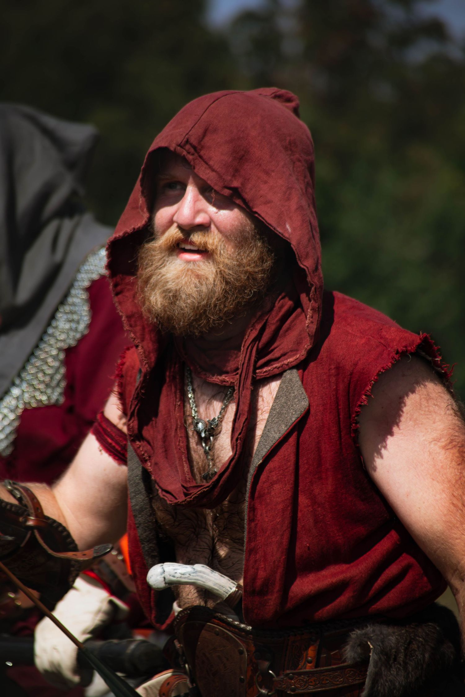 Medieval reenactor in red hooded cloak and leather armor with beard in natural outdoor setting.