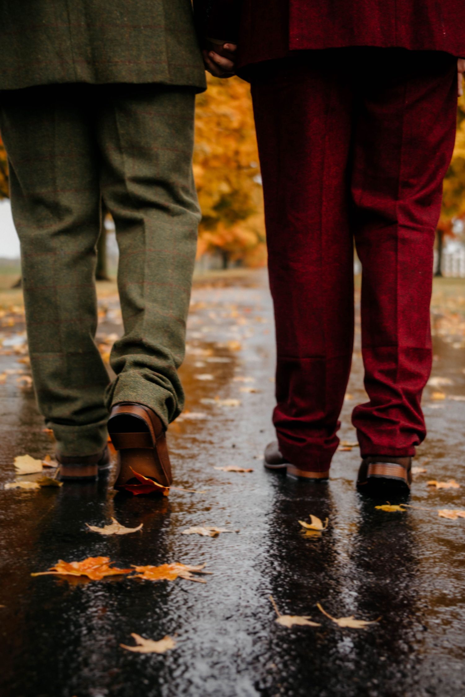 Two pairs of legs wearing contrasting gray and burgundy pants stand on wet pavement with autumn leaves scattered around.