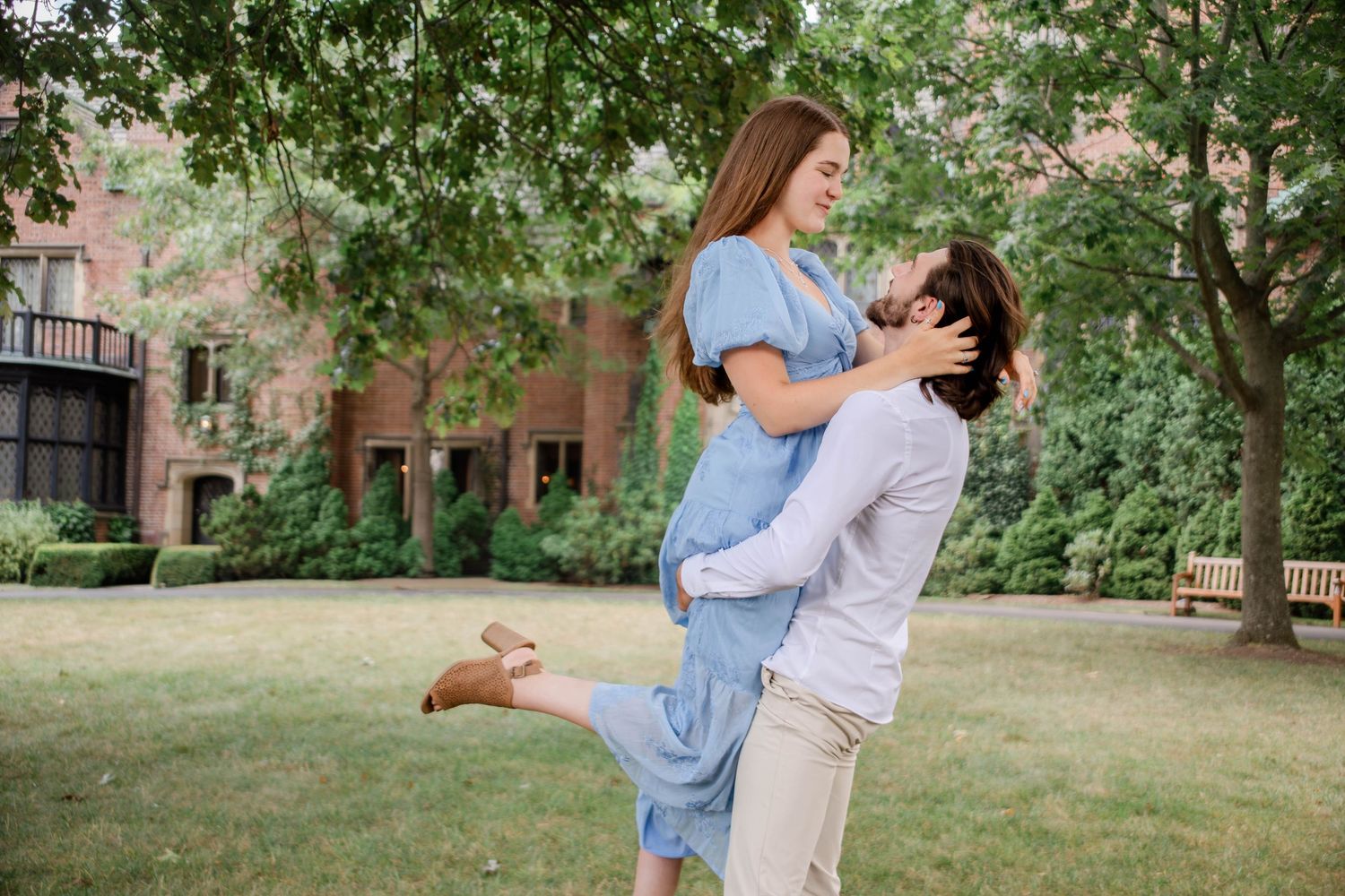 Romantic couple shares tender moments in an outdoor park setting with brick buildings in background.