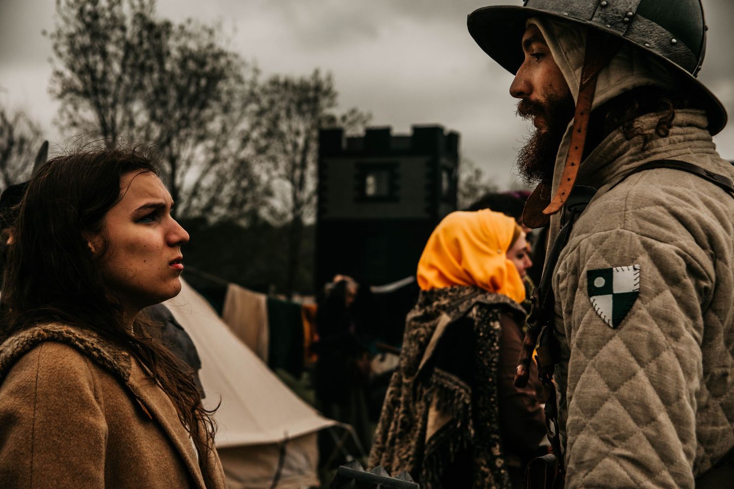 Two people having an intense conversation outdoors with a tent and dark cloudy sky in the background.