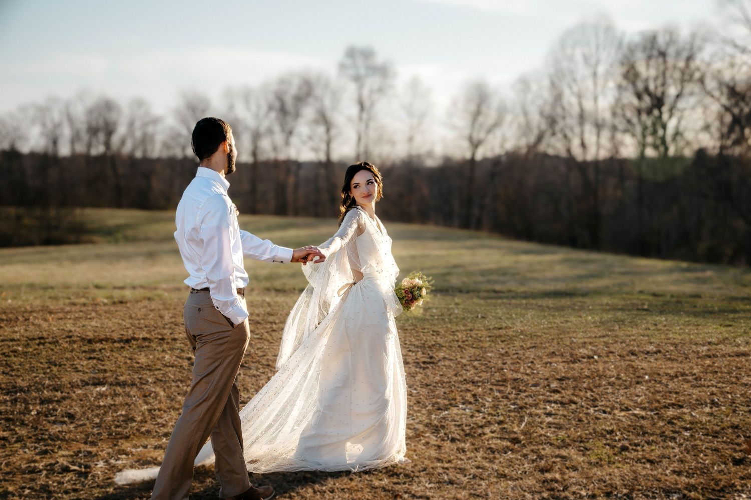 A couple in wedding attire holds hands in a golden field at sunset with bare trees in the background.