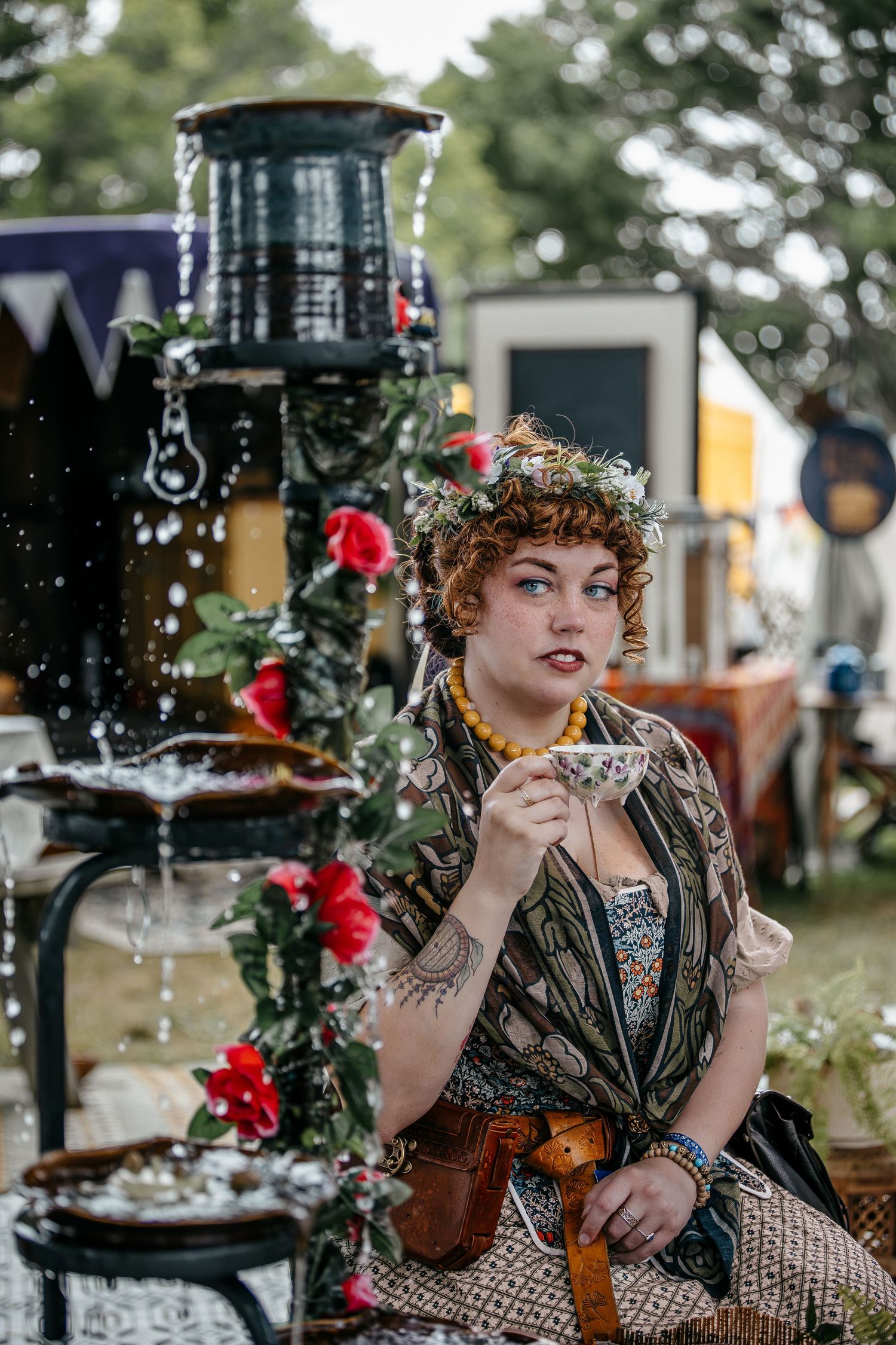 A woman in bohemian costume with floral crown sits near a decorative fountain with red roses at an outdoor festival.
