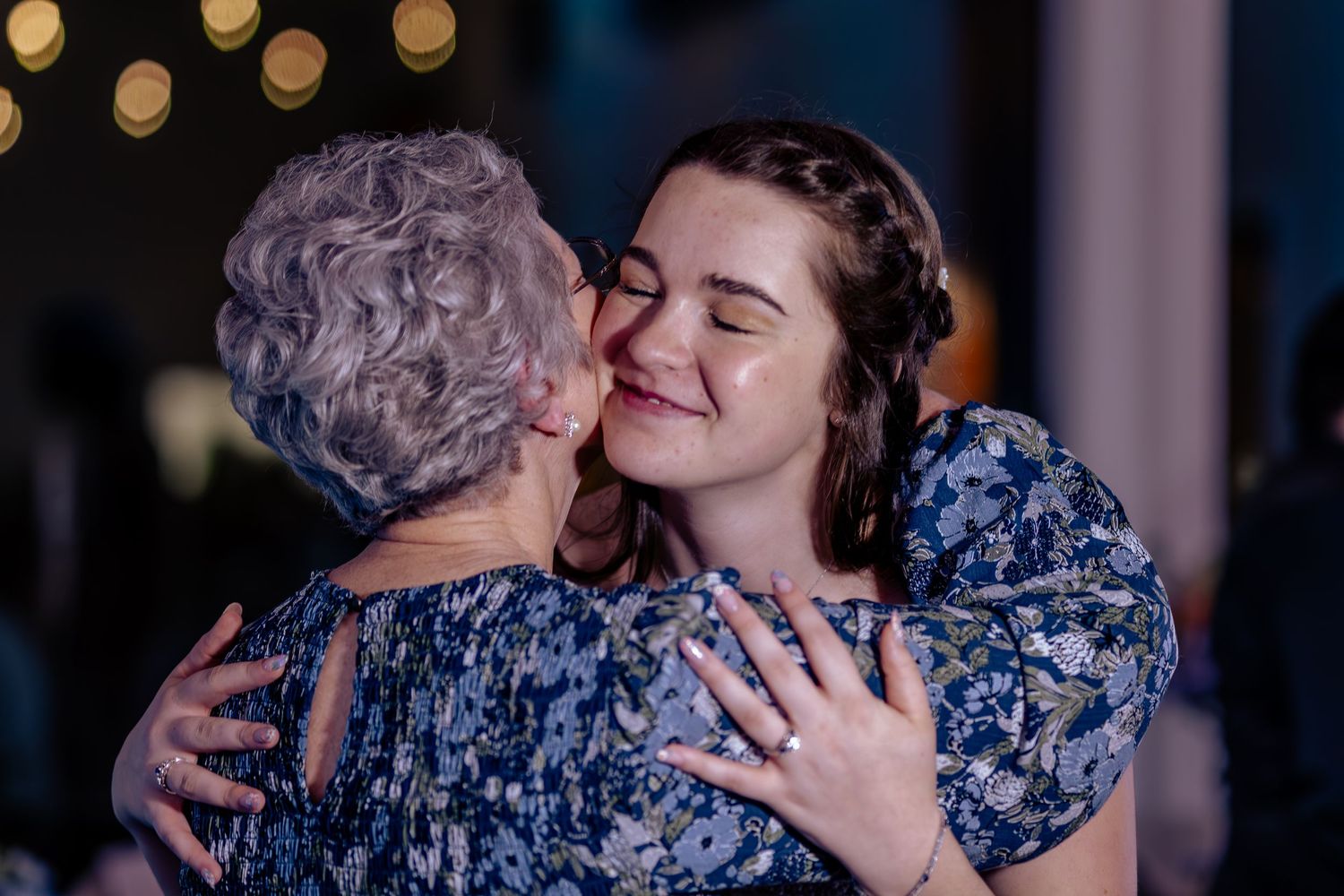 Two women share a warm embrace and kiss at a formal event, one elderly with gray hair, one younger in a floral dress.