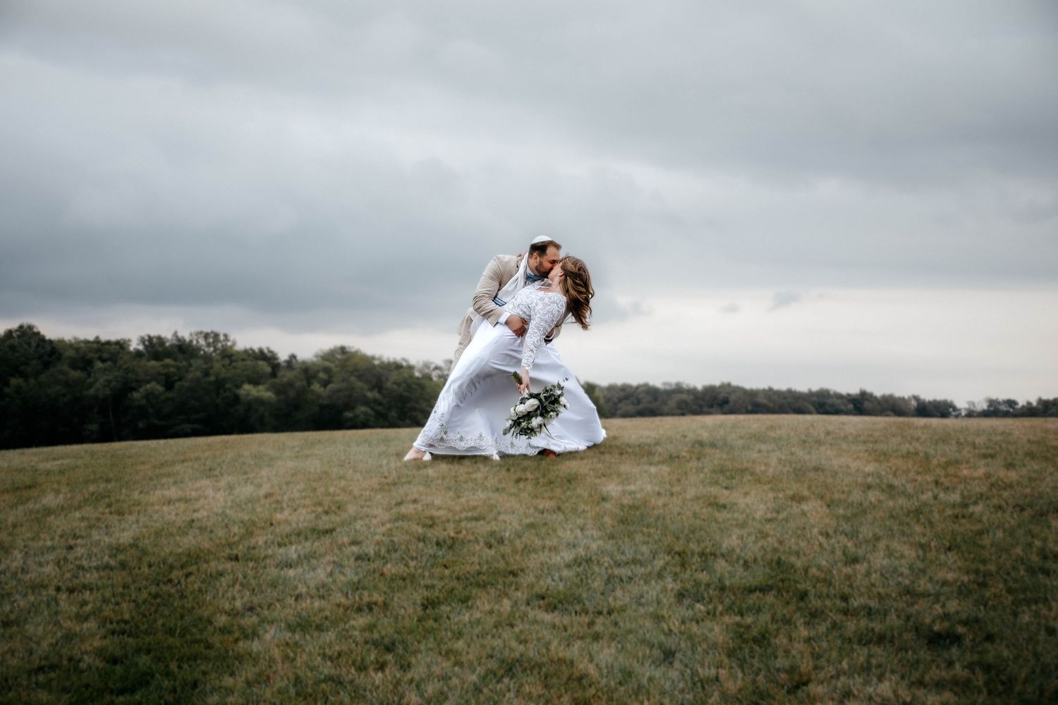 A couple shares a romantic dip kiss in a green field on their wedding day under a cloudy sky.