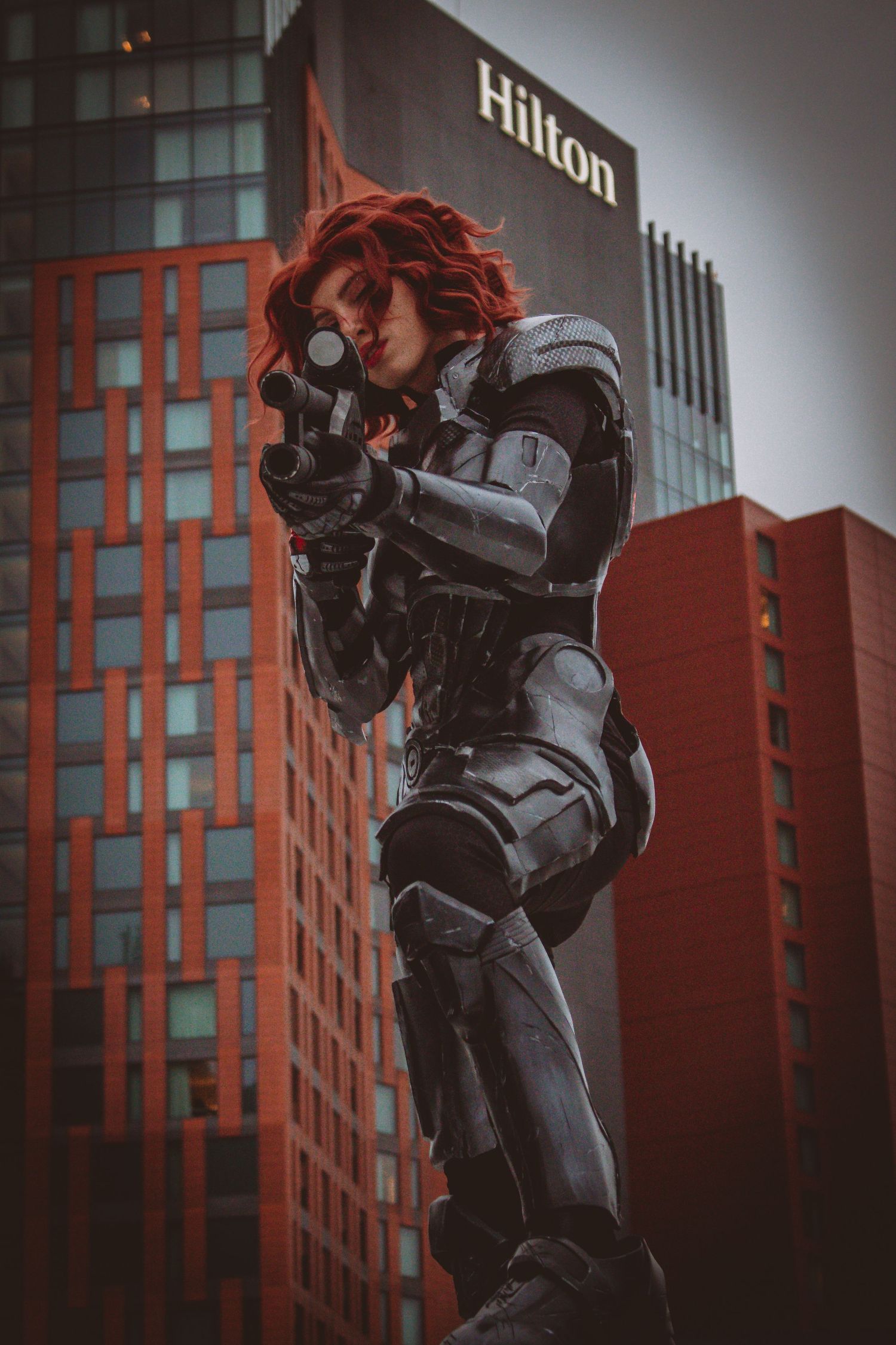 A cosplayer in a black combat suit poses with a weapon against the backdrop of a red Hilton hotel building.