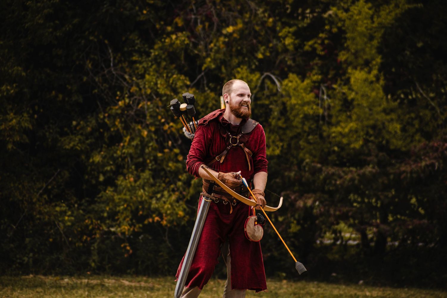 A person in a red shirt practices archery in a wooded outdoor setting with autumn foliage in the background.