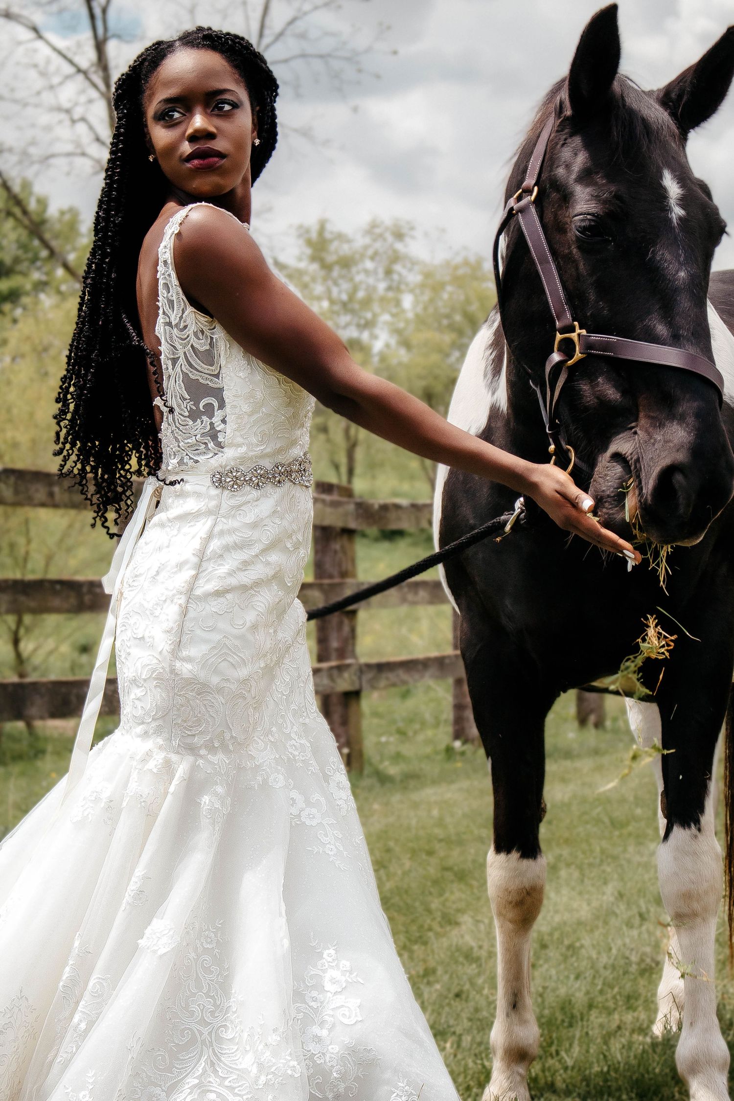 A beautiful bride in a white lace dress poses with a black and white horse in a pastoral outdoor setting.