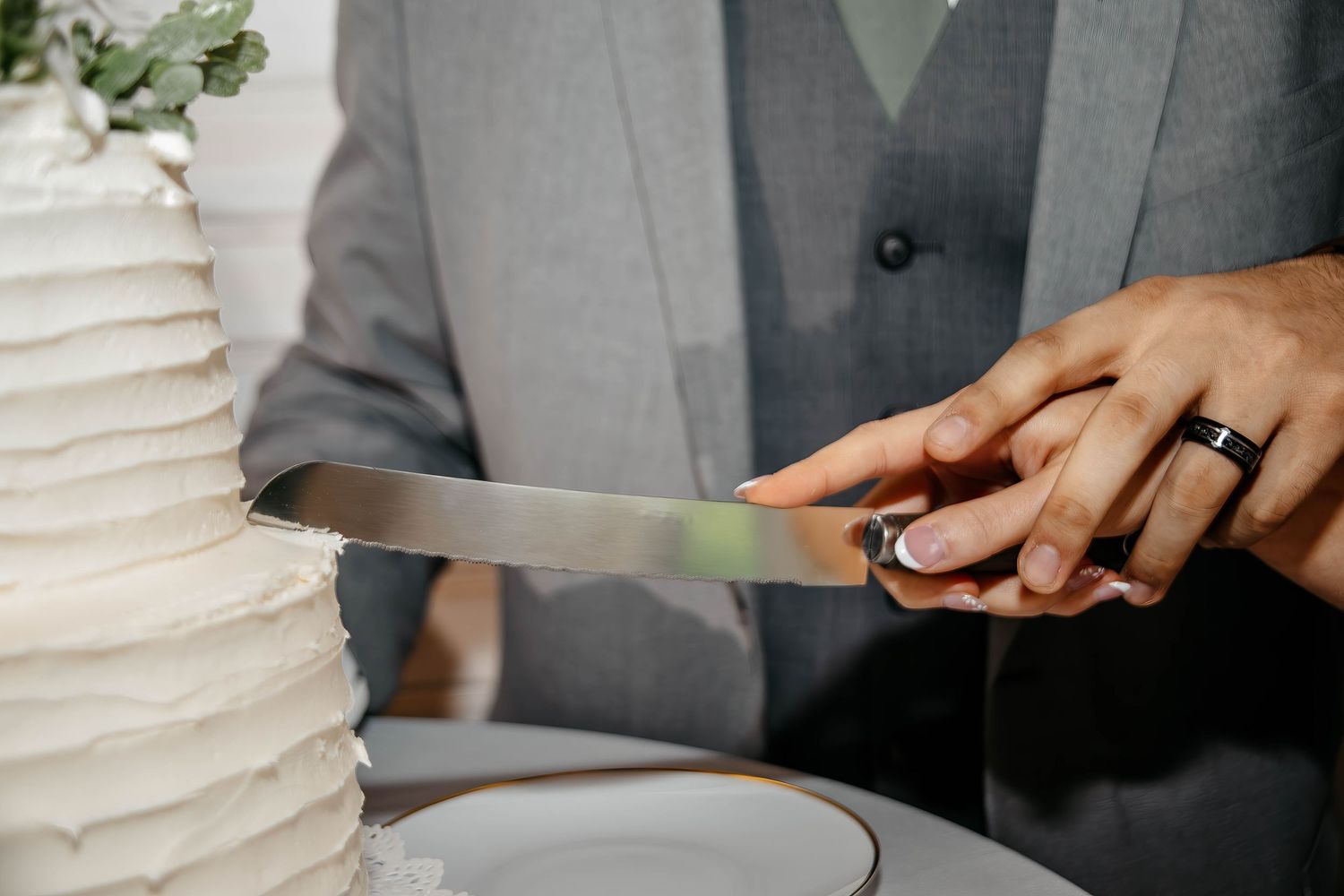 A wedding cake being cut by hands wearing formal attire during an elegant reception celebration.
