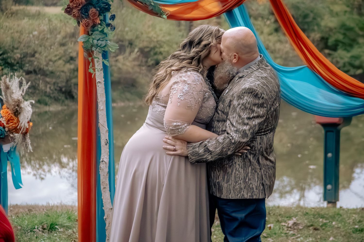 A couple shares an intimate moment in front of a lake with a decorative orange and teal wedding arch.