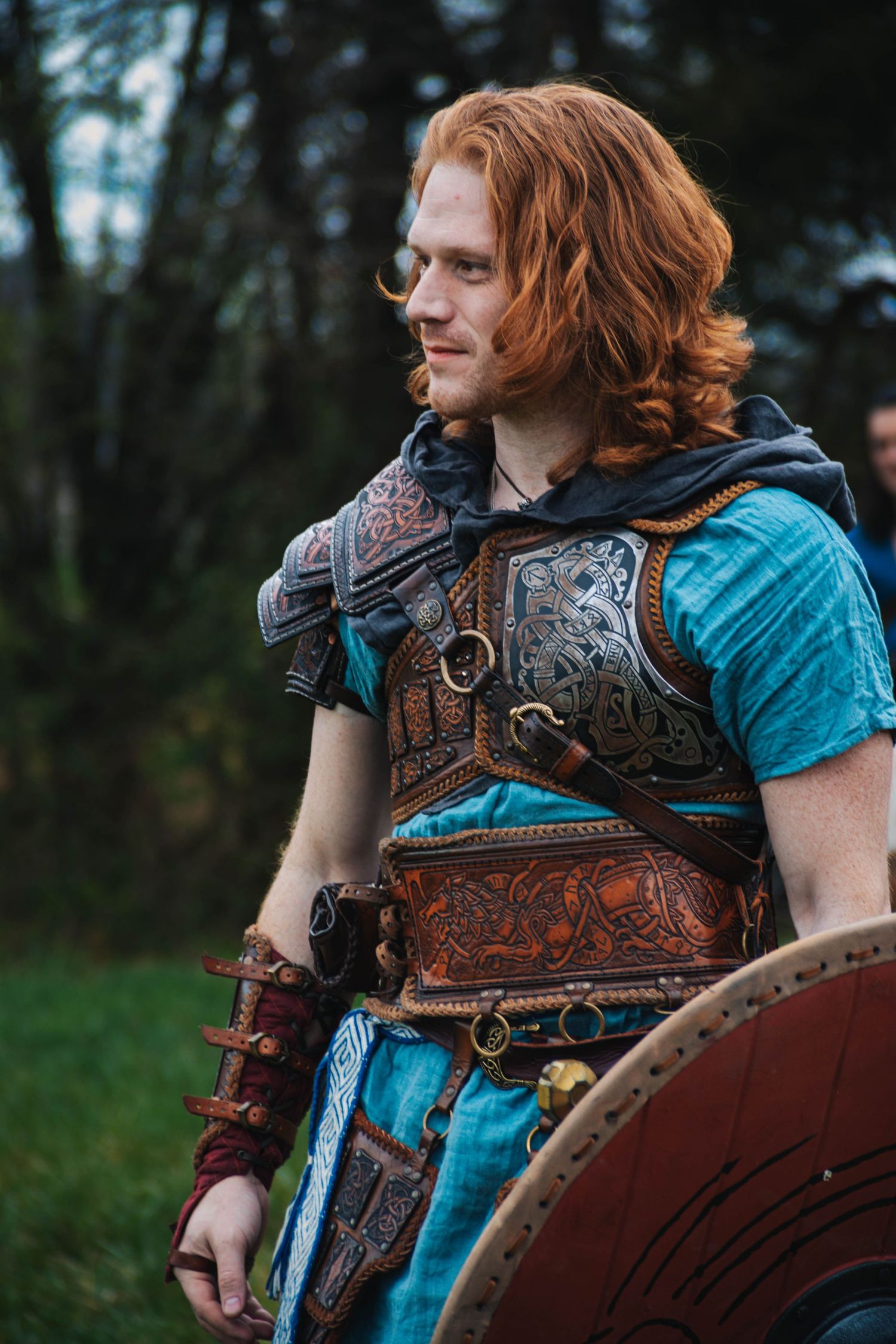 Medieval warrior wearing ornate leather armor with detailed tooling and a turquoise tunic in a forest setting.