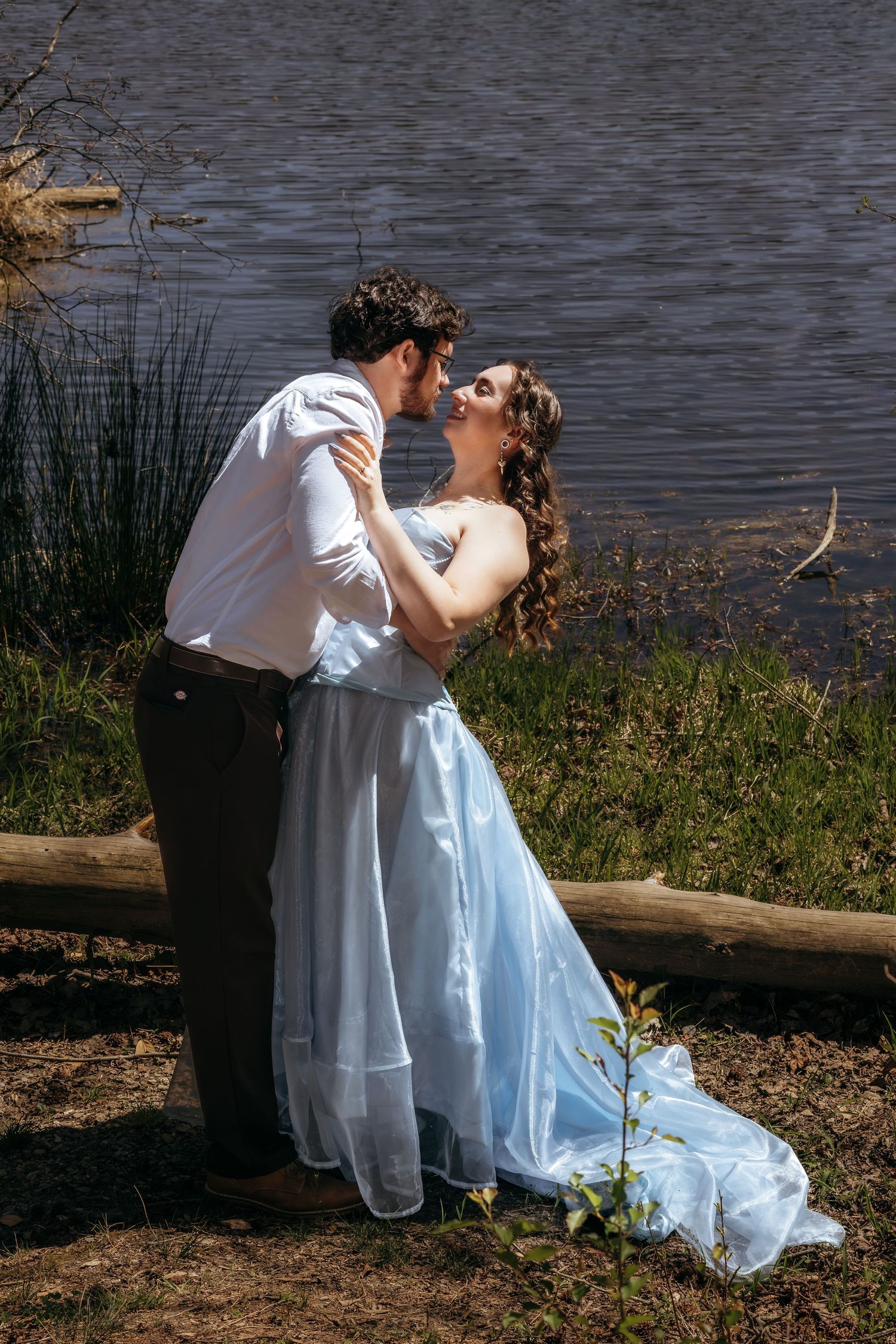 Couple in wedding attire sharing a romantic moment by a lake, bride in light blue gown embracing groom in white shirt.