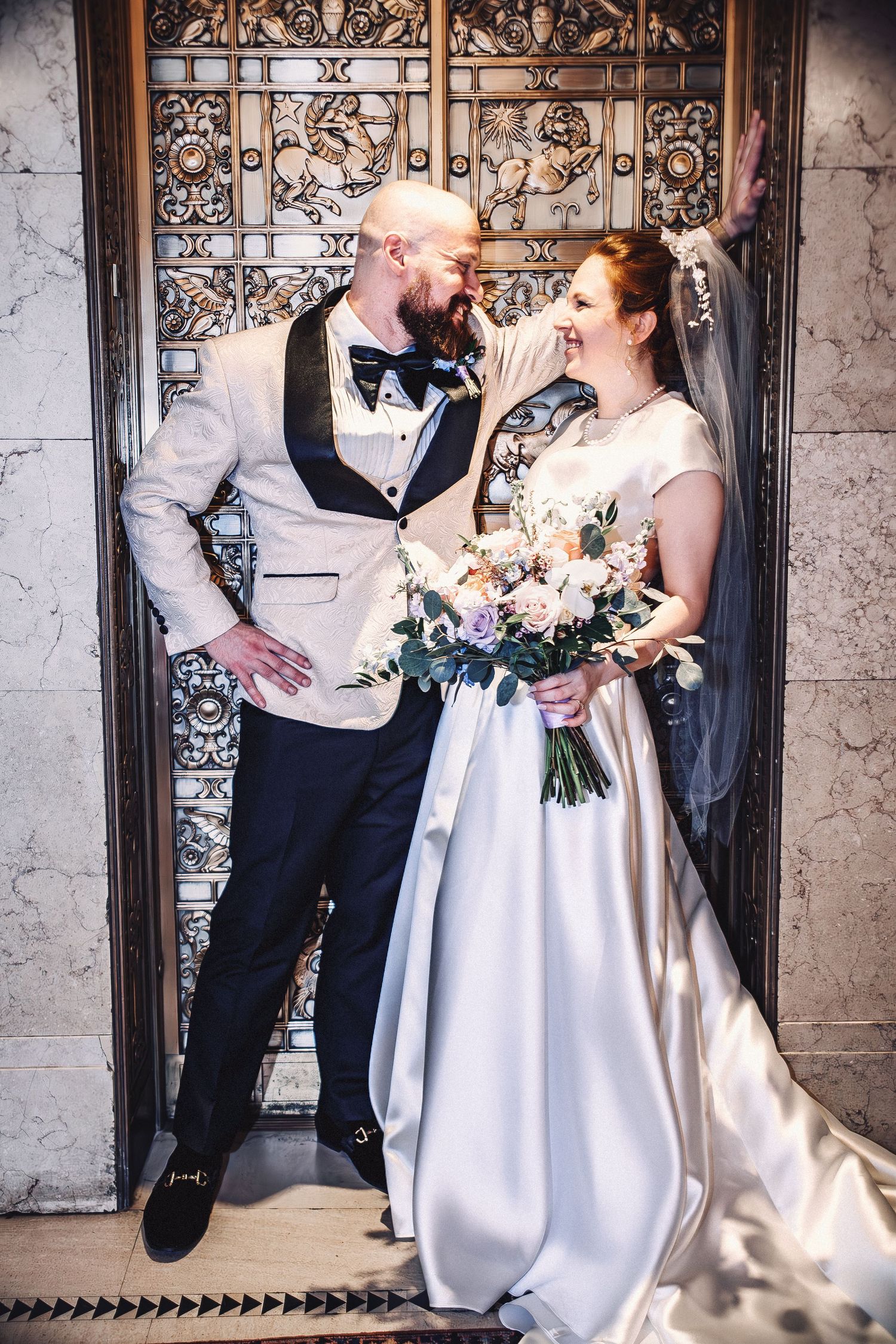 Bride and groom pose elegantly in front of ornate doors, bride holding a pastel bouquet in a satin gown.