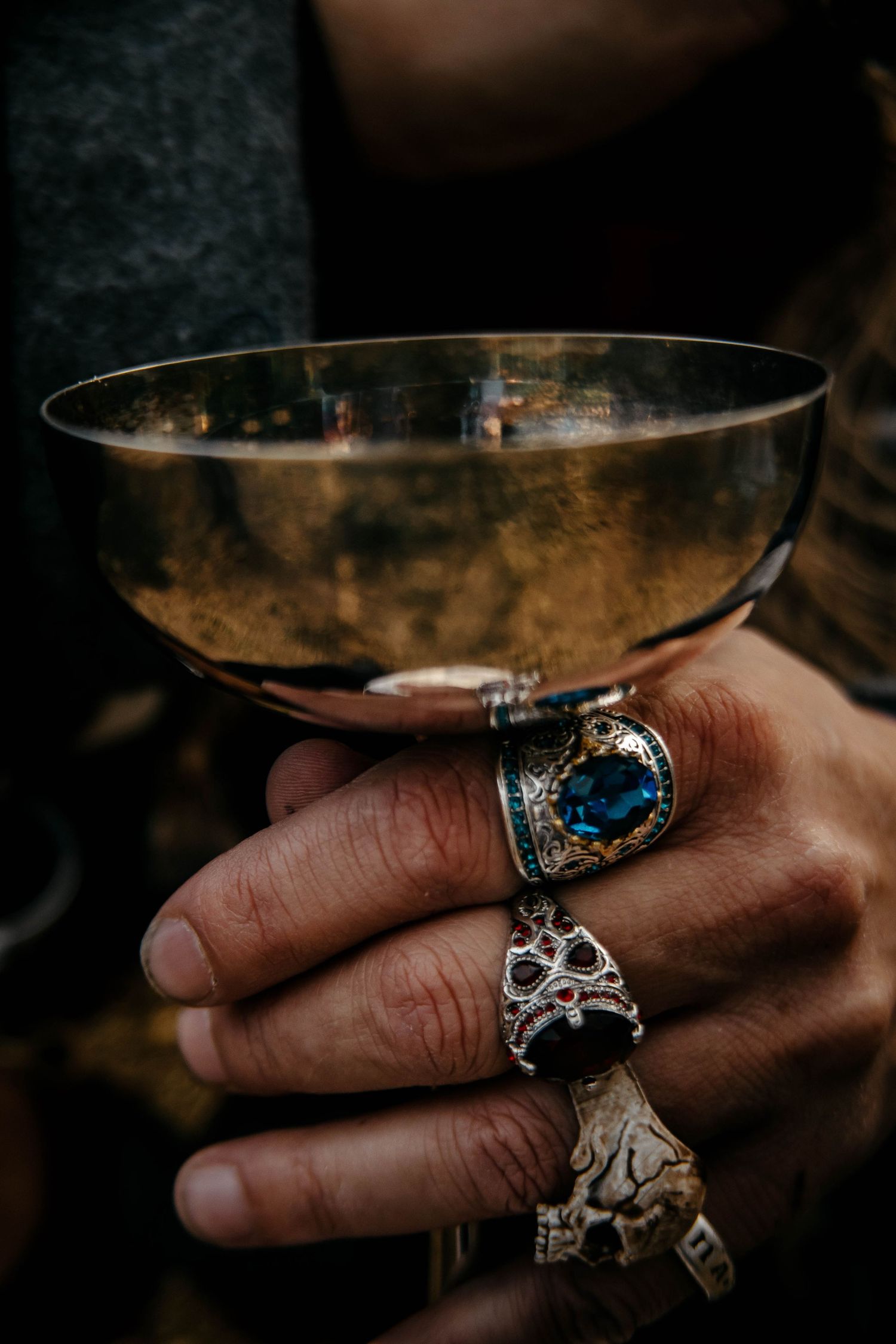 A hand wearing ornate silver and blue gemstone rings holds a glass of wine in a dark moody setting.