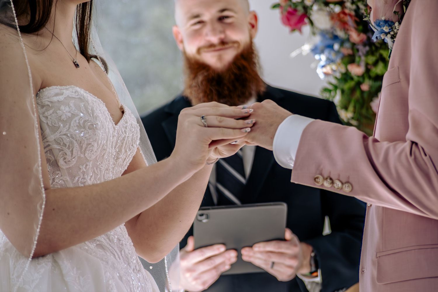 A group of wedding guests in blush pink dresses read from papers during an outdoor ceremony.
