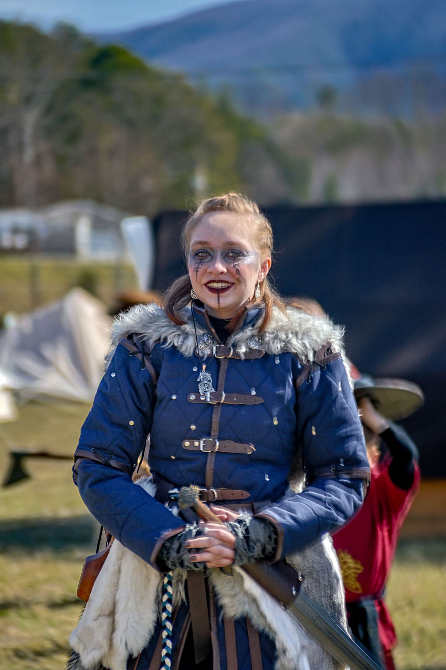 A medieval reenactor in a blue fur-trimmed coat and face paint stands outdoors at a historical event.