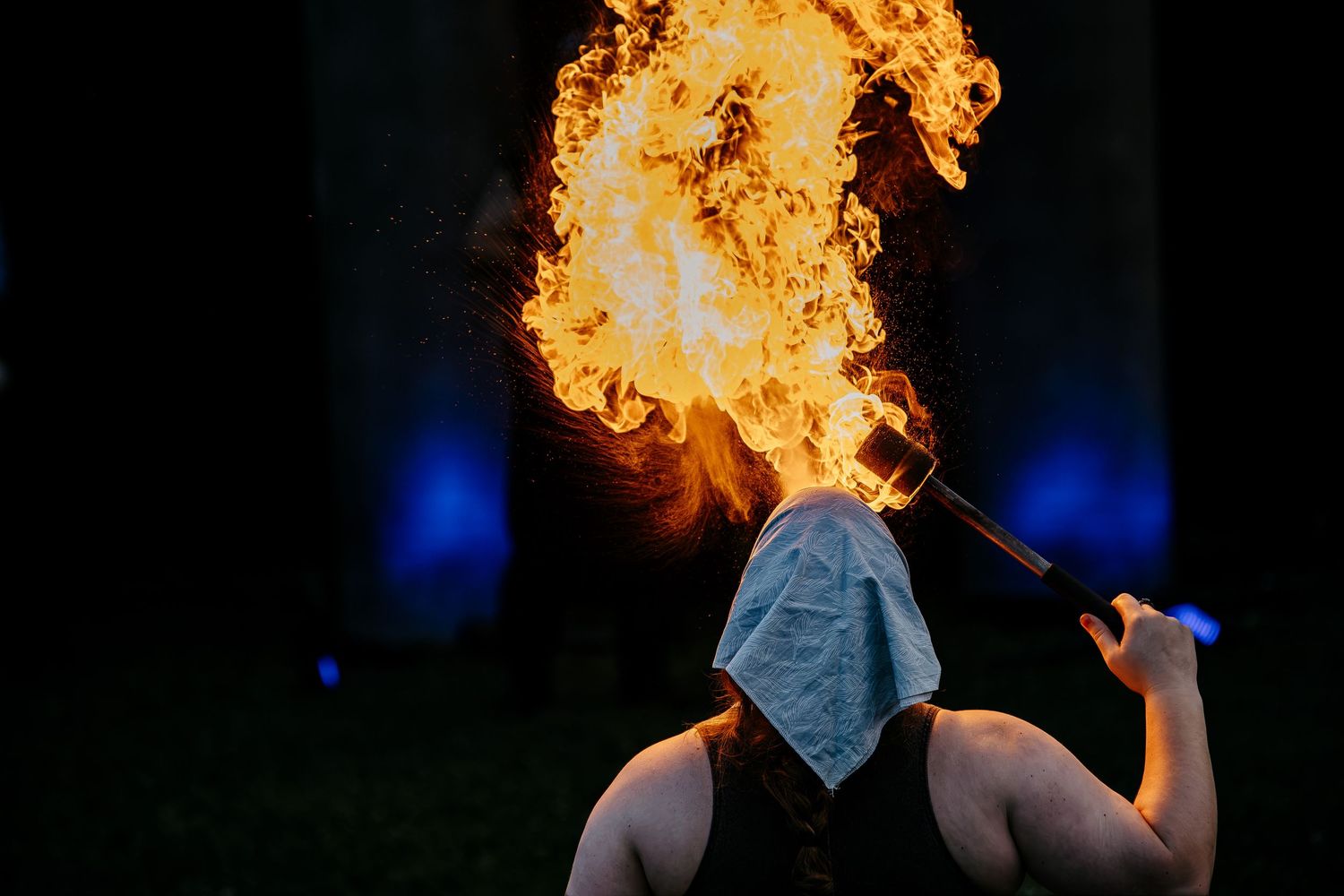 Fire breather performing a dramatic flame stunt outdoors at night, wearing a white head covering and sleeveless top.