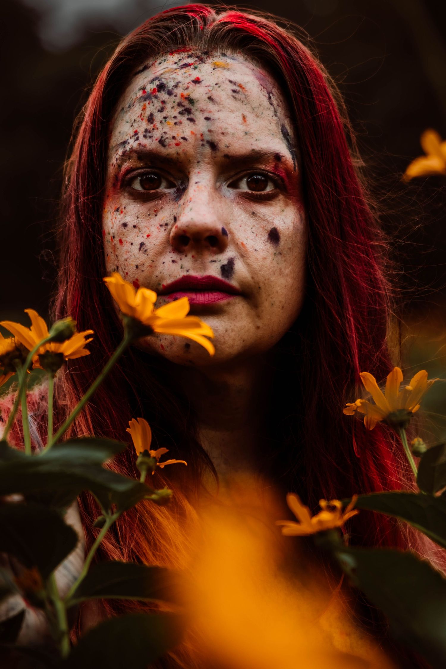 Artistic portrait with vibrant red hair and speckled face makeup amidst blooming orange flowers in moody lighting.