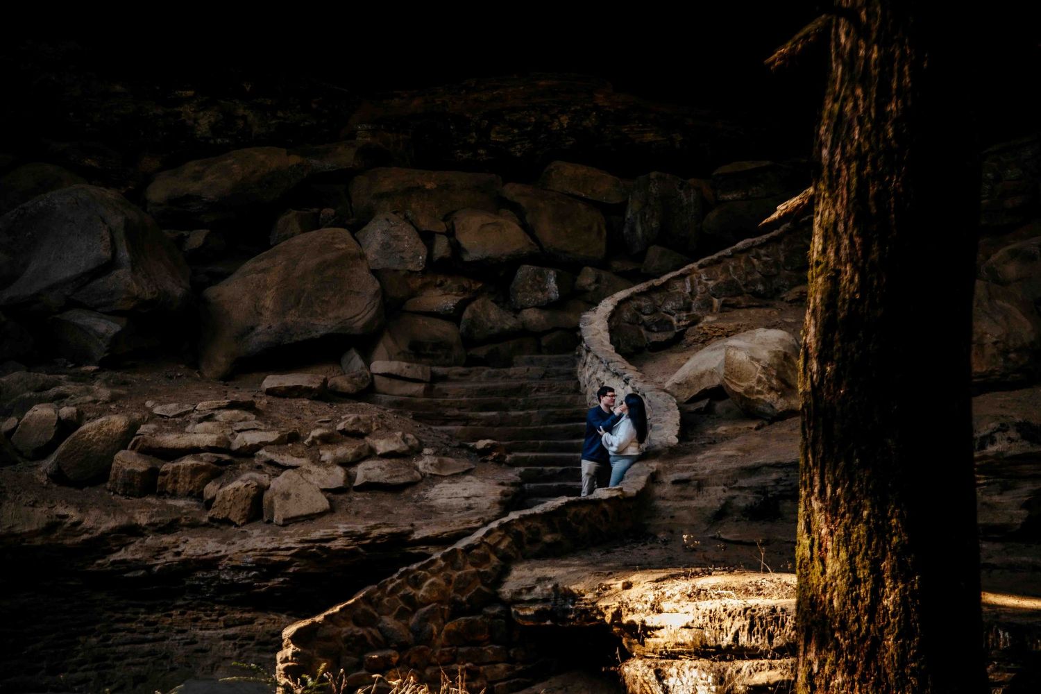 Couple embracing on rocky riverbank surrounded by boulders and trees in a dramatic dark outdoor engagement photo.