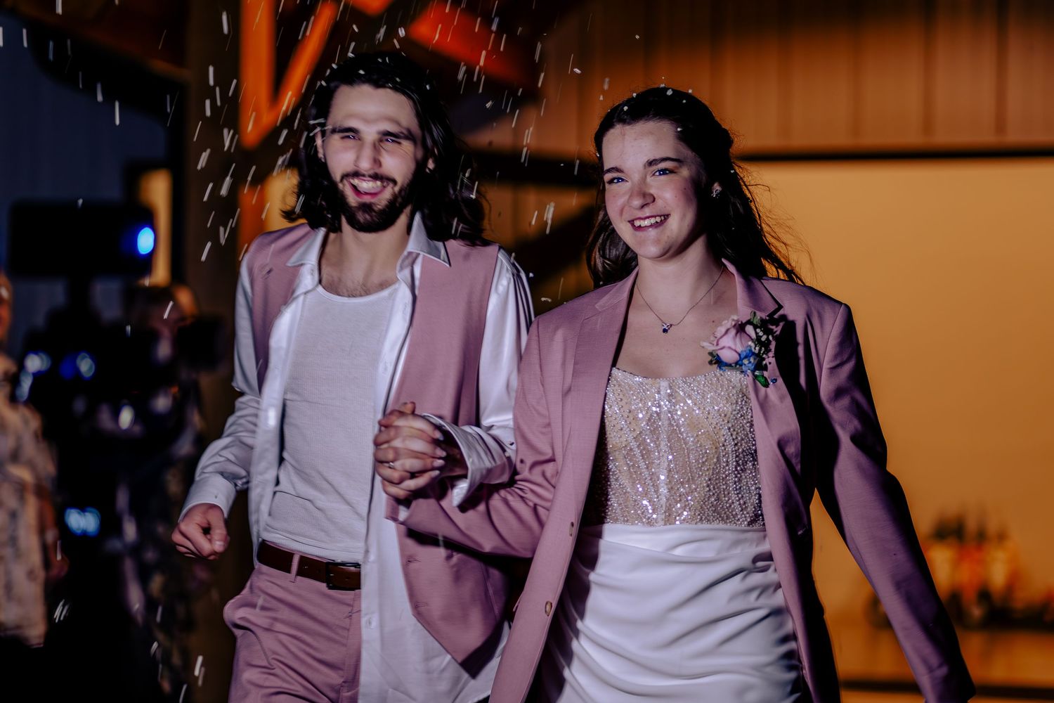 Happy newlyweds in matching pink blazers walk together, smiling, with confetti falling at their wedding reception.