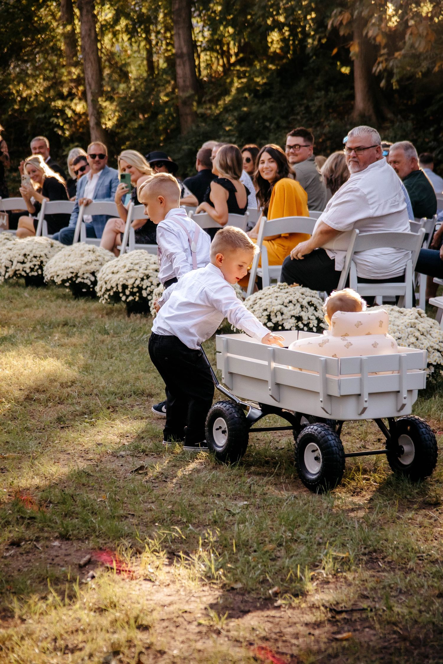 A young ring bearer pushes a white wagon carrying a flower girl down the aisle at an outdoor wedding ceremony.