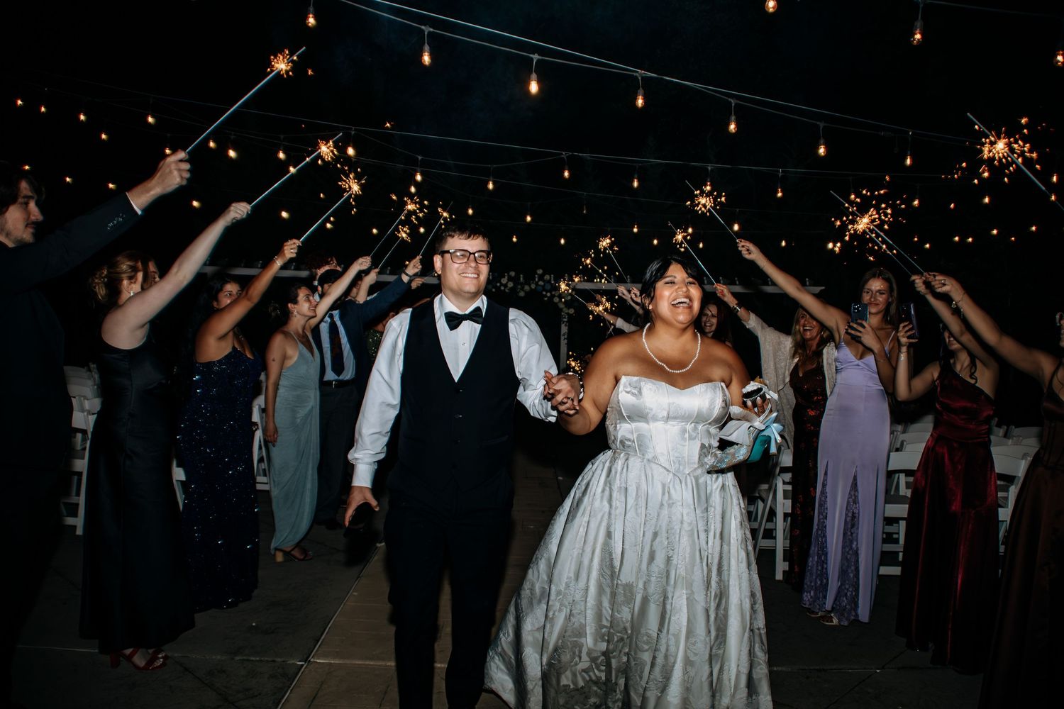 Bride and groom walk through sparkler tunnel with wedding guests at night outdoor reception celebration.