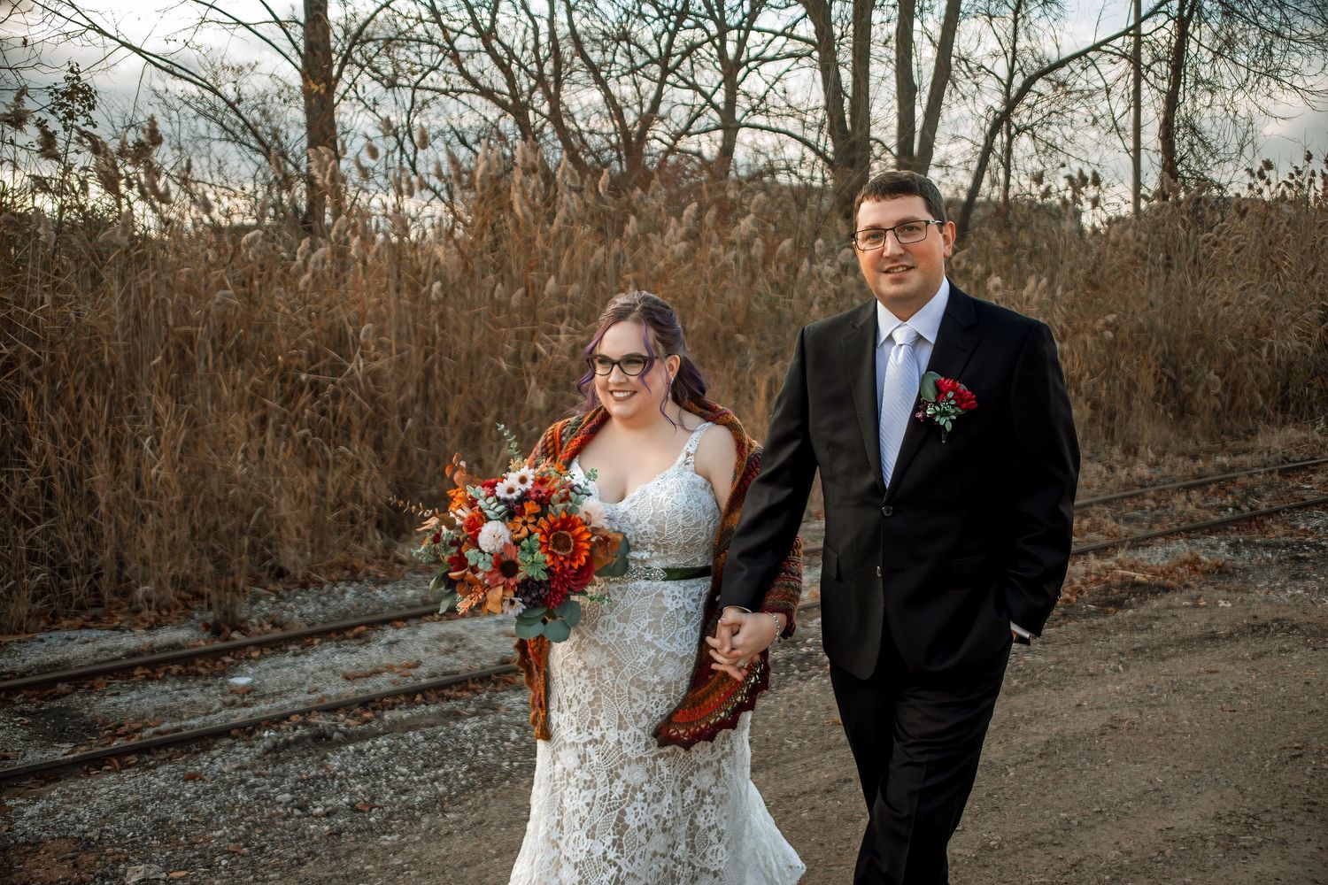 A couple in wedding attire walks along a rustic autumn path holding a vibrant orange and red floral bouquet.