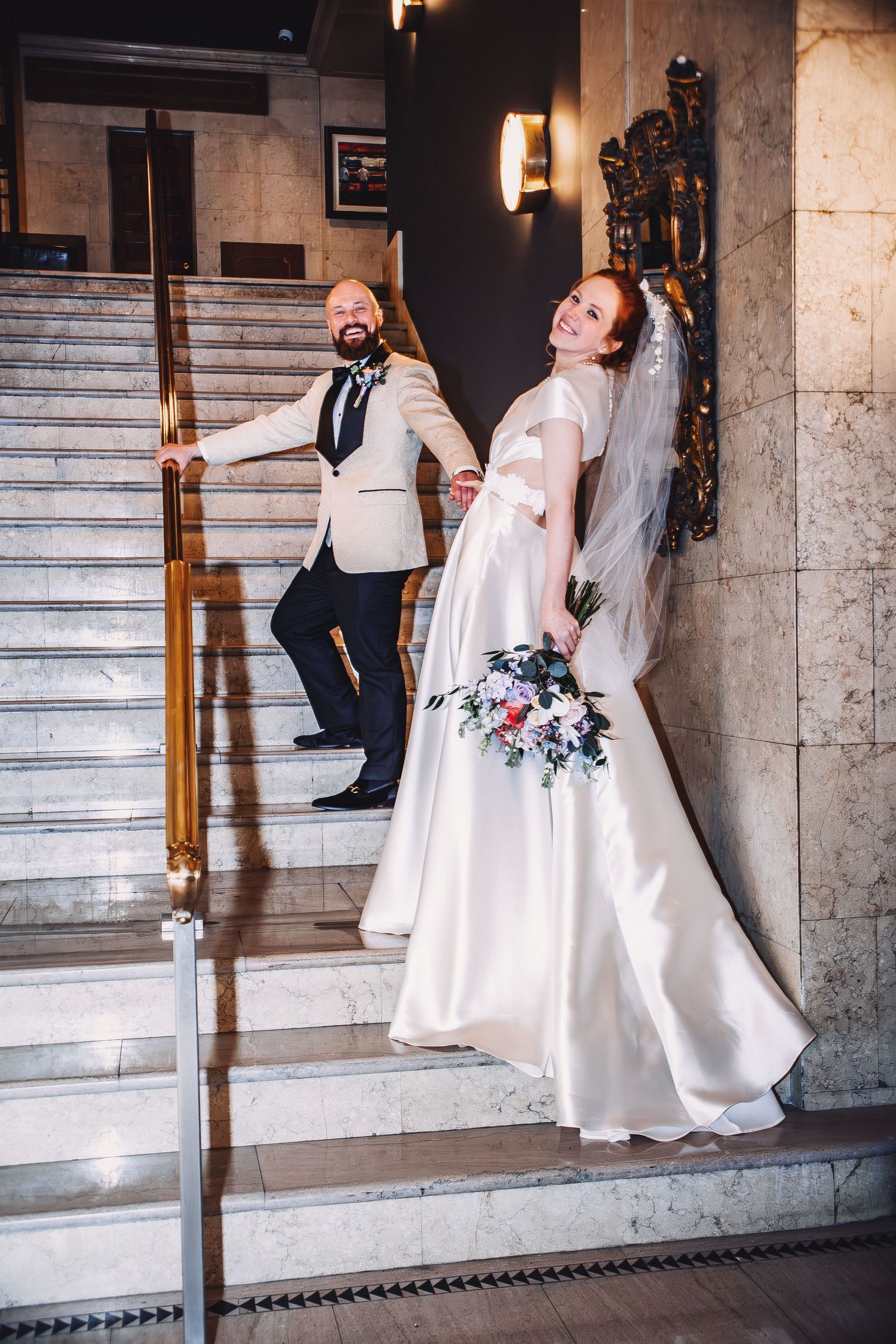 Bride in white gown holding bouquet and groom in beige tuxedo posing on marble staircase in elegant venue.