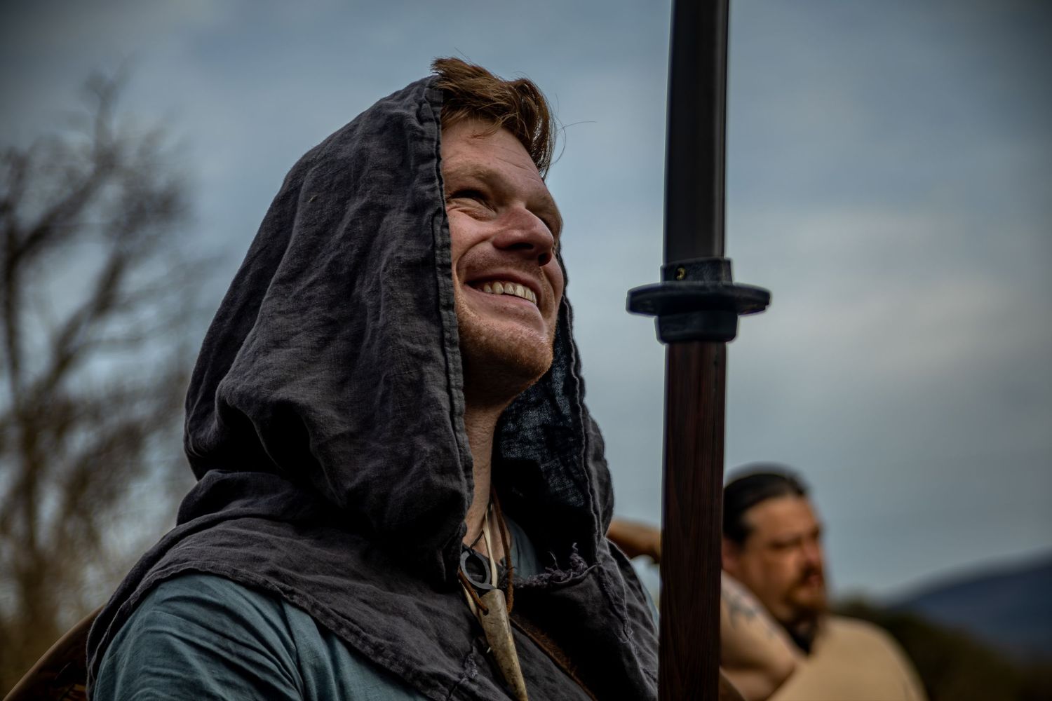 A person in a black hooded jacket stands against a moody sky background.