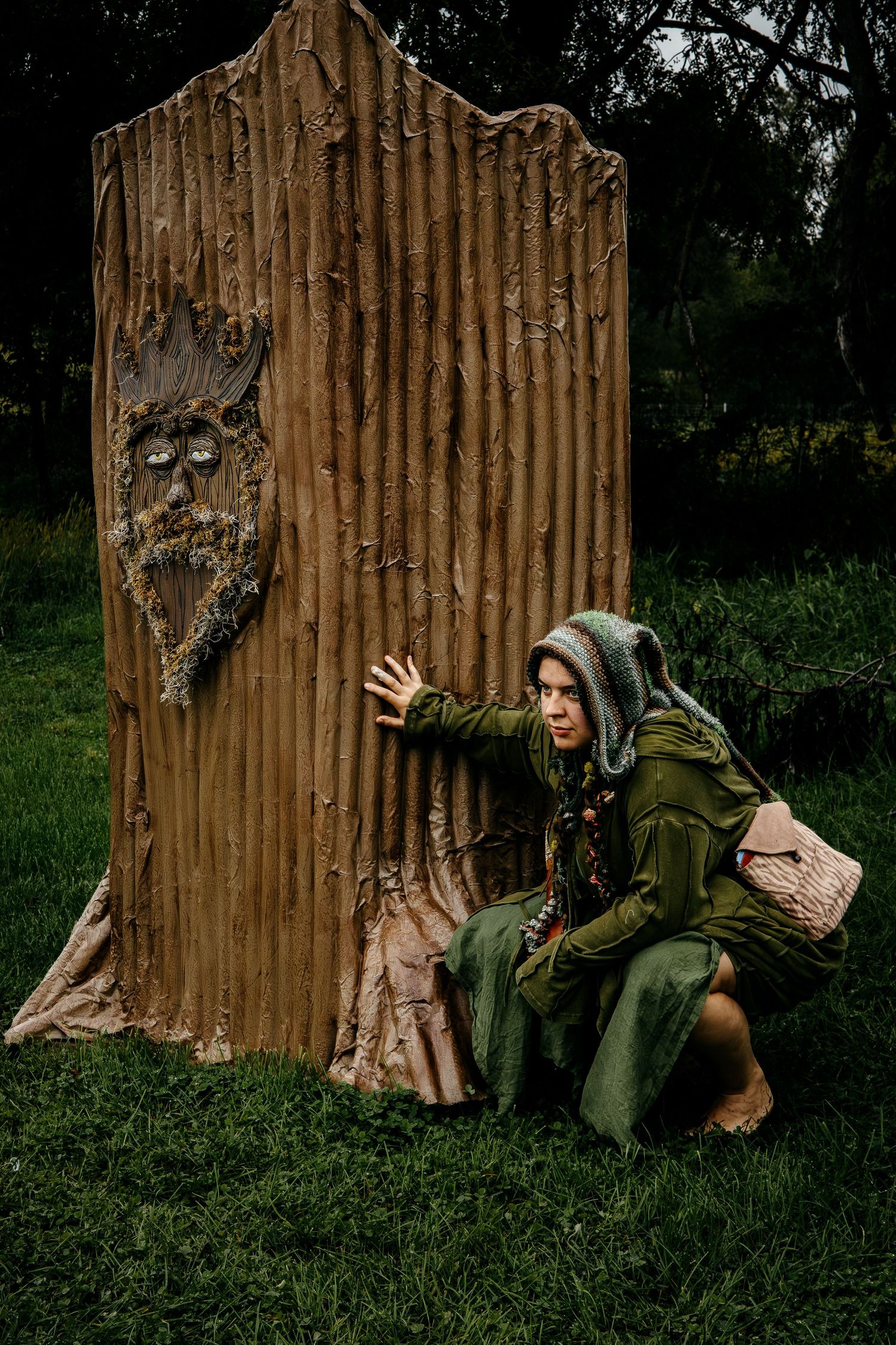 Woman in green hooded cloak crouching beside a carved wooden tree stump with a face in a forest setting.